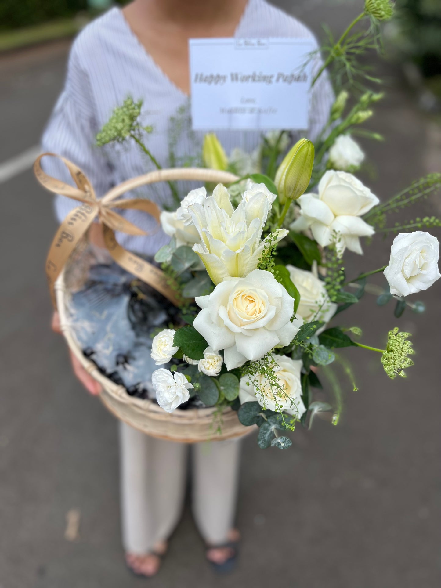 "Keranjang rotan elegan berisi anggur segar, bunga lili, dan mawar putih yang indah, disusun dengan rapi untuk dijadikan hamper."