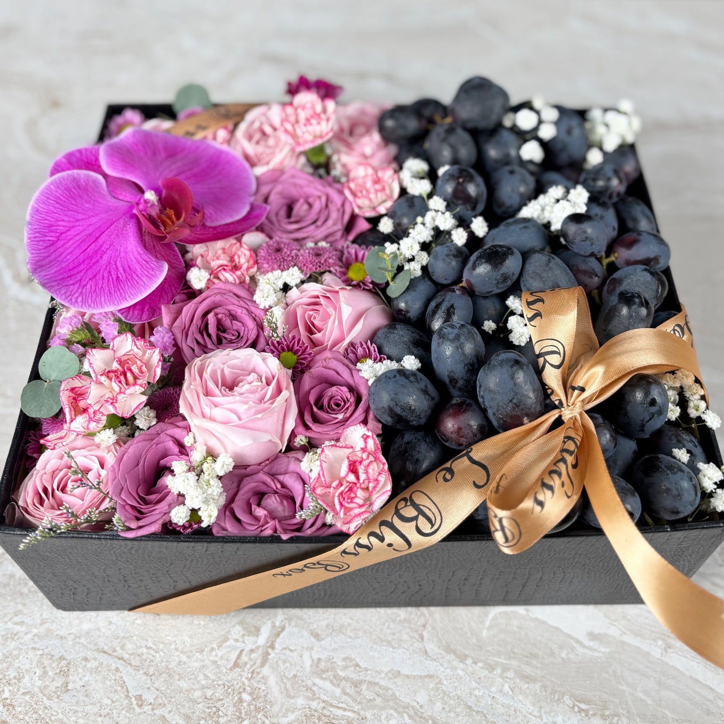Box of flowers and grapes with a gold ribbon on a light background. Pink Lilac Sonata Autumn Grape Box dari Le Bliss Bouquet. Kado Hari Ibu elegan dengan bunga & buah premium. Pengiriman Surabaya, Jakarta & Cirebon. Soft, sweet & luxurious. Kado Anggur dan anggrek untuk Hari Ibu