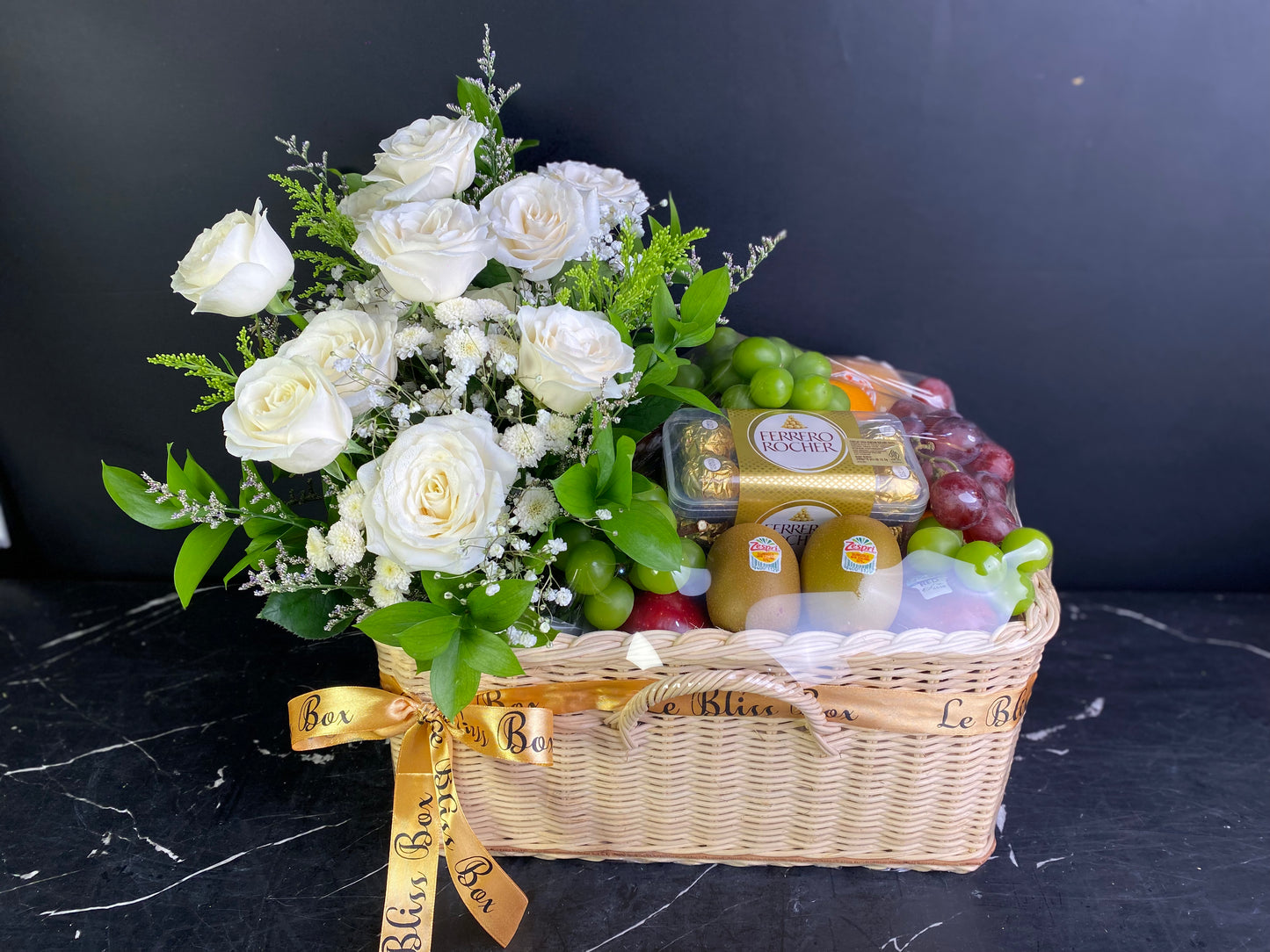 Gift basket with flowers, fruits, and a bottle on a dark surface. Fruit basket with white roses and a gold ribbon on a dark background. Florist Le Bliss Box parcel buah surabaya, parcel buah jakarta, coklat hamper buah surabaya