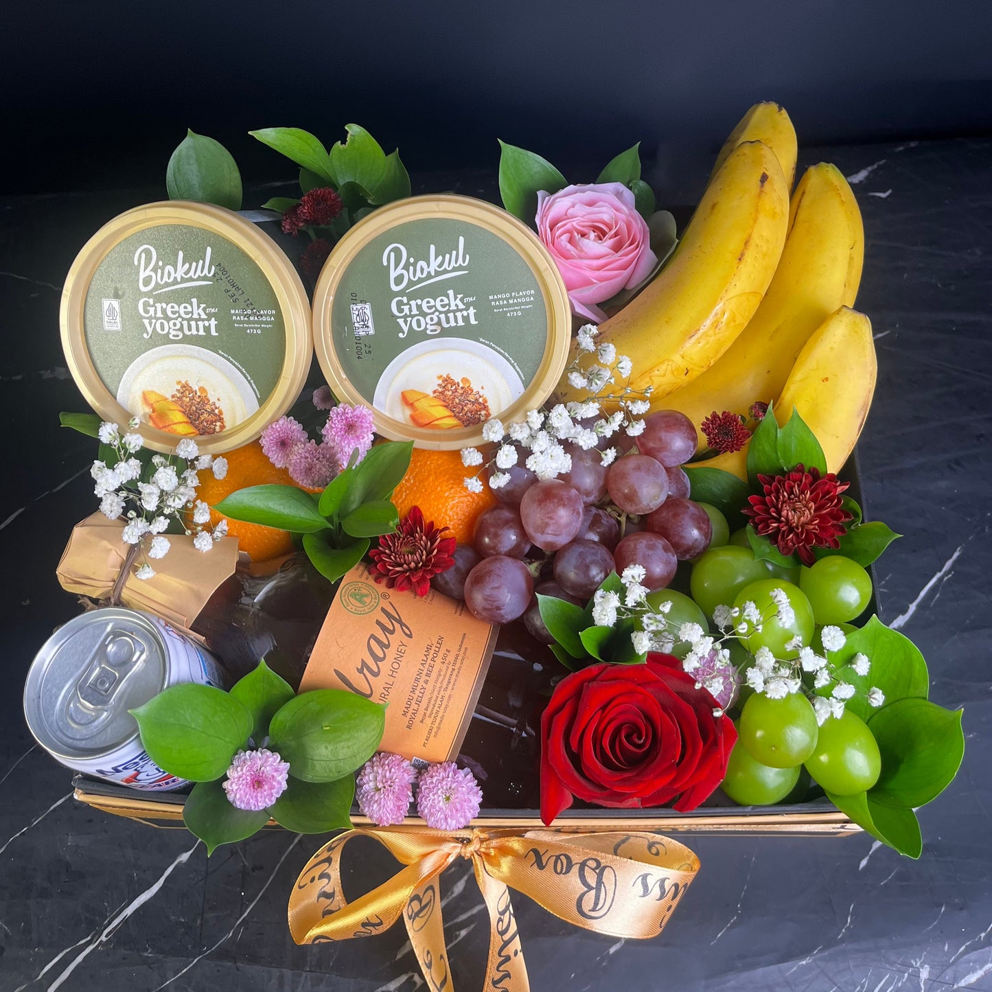 Gourmet gift basket with yogurt, fruit, and flowers on a dark surface. Fruit and yogurt basket with a red rose on a dark background. yogurt, honey, bear brand, banana, orange fruit hamper surabaya, jakarta, cirebon