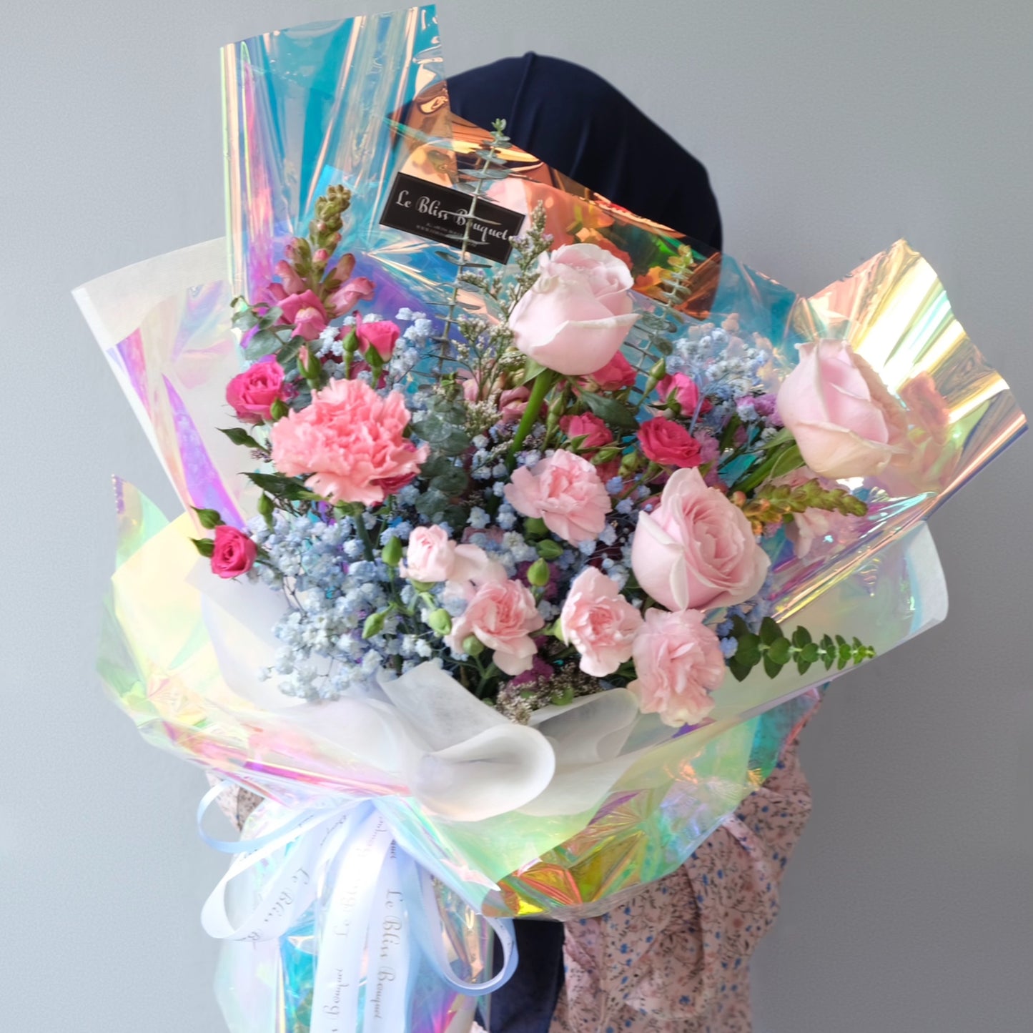 Person holding a bouquet of flowers wrapped in iridescent cellophane against a plain background. Le Bliss Bouquet’s Pink Hologram Bouquet — trendy pink roses wrapped in holographic paper. Same-day delivery to Jabodetabek, Surabaya & Cirebon.