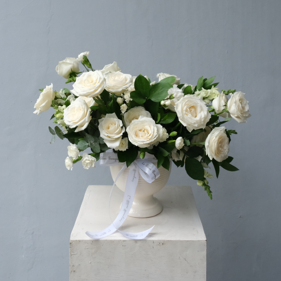 A floral arrangement featuring white avalanche roses in a white vase, placed on a white pedestal against a grey background. Rangkaian mawar avalanche putih dalam vas kaca bening, disusun elegan dan minimalis — Pure White Avalanche Rose Vase dari Le Bliss. Surabaya florist