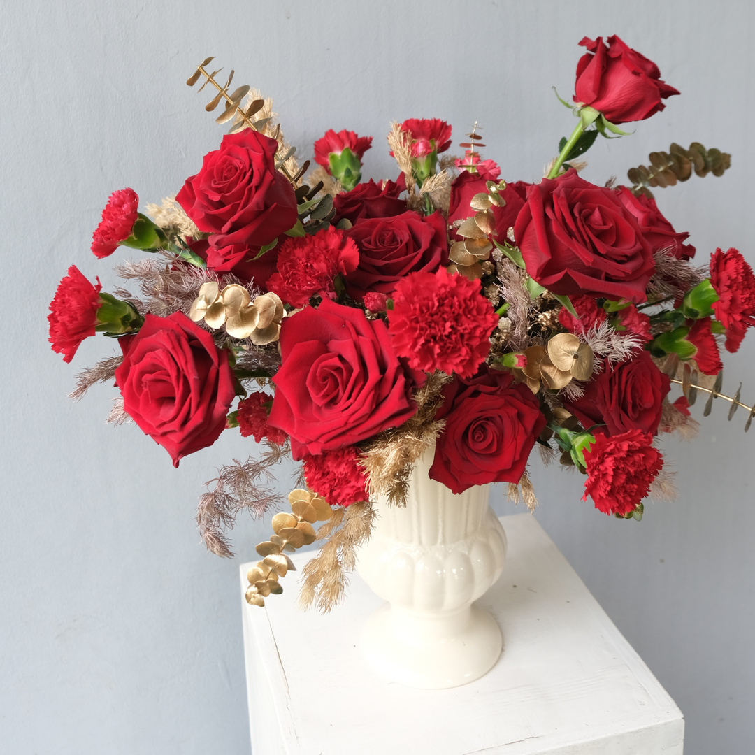 A vibrant floral arrangement featuring red roses and carnations in a white vase with gold decorations. Rangkaian mawar merah Ecuador dalam vas kaca elegan, simbol cinta dan penghormatan — Regal Explorer Rose Vase dari Le Bliss. florist surabaya, toko bunga surabaya