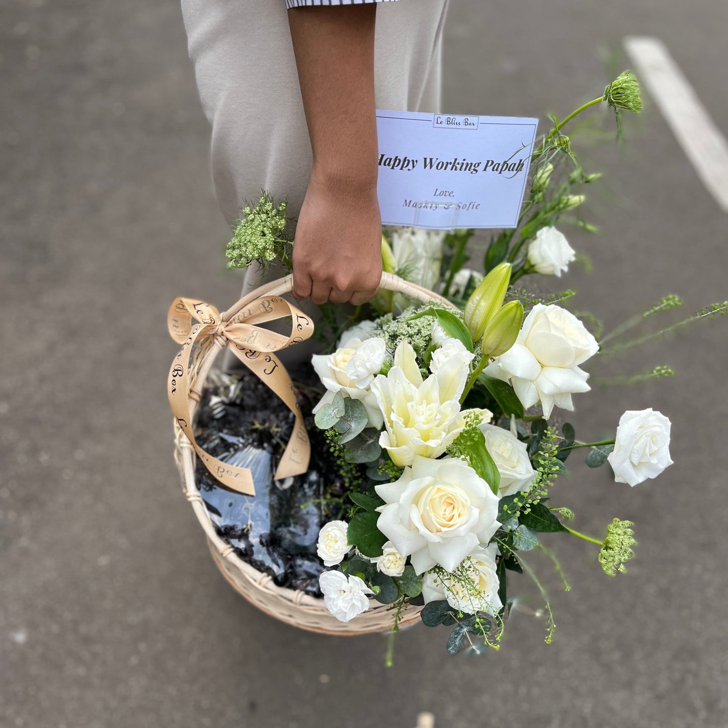 Keranjang rotan elegan berisi anggur segar, bunga lili, dan mawar putih yang indah, disusun dengan rapi untuk dijadikan hamper.