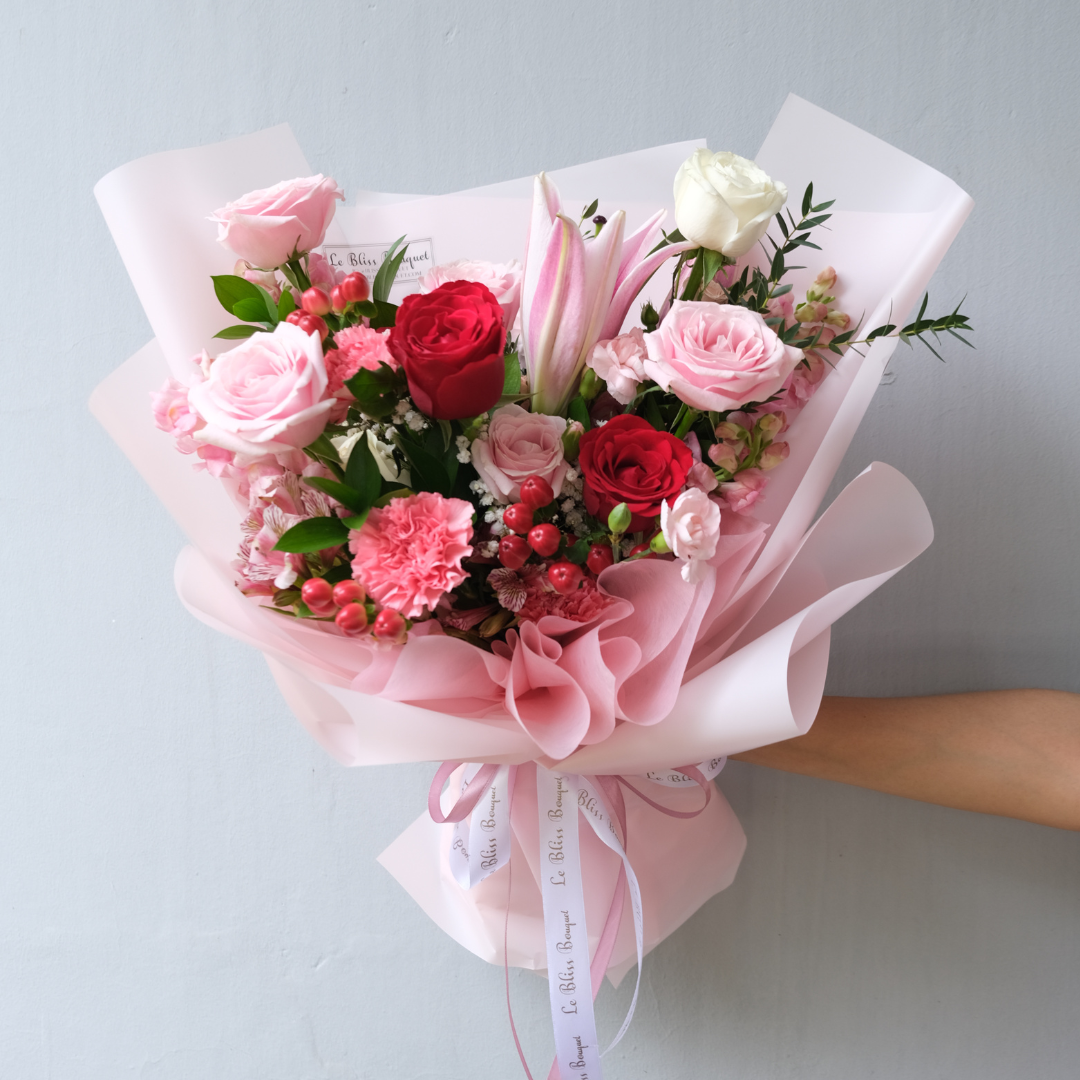 A medium bouquet with a mix of red and pink flowers, including roses and lilies, wrapped in pink paper with a white ribbon. Le Bliss Bouquet surabaya, toko bunga surabaya, florist surabaya, bouquet bunga surabaya