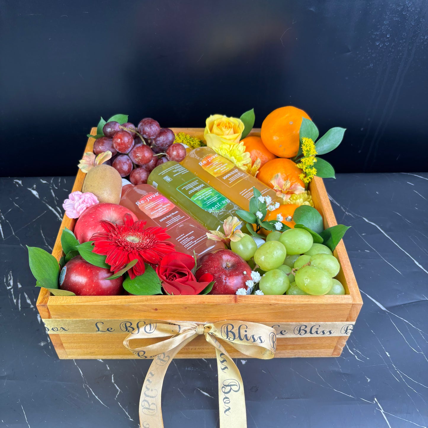 A wooden fruit box containing an assortment of fresh fruits such as apples, oranges, grapes, and bananas, along with two bottles of juice and decorative flowers. rejuve juice fruit hamper jakarta, surabaya, cirebon. parcel buah dengan juice revue