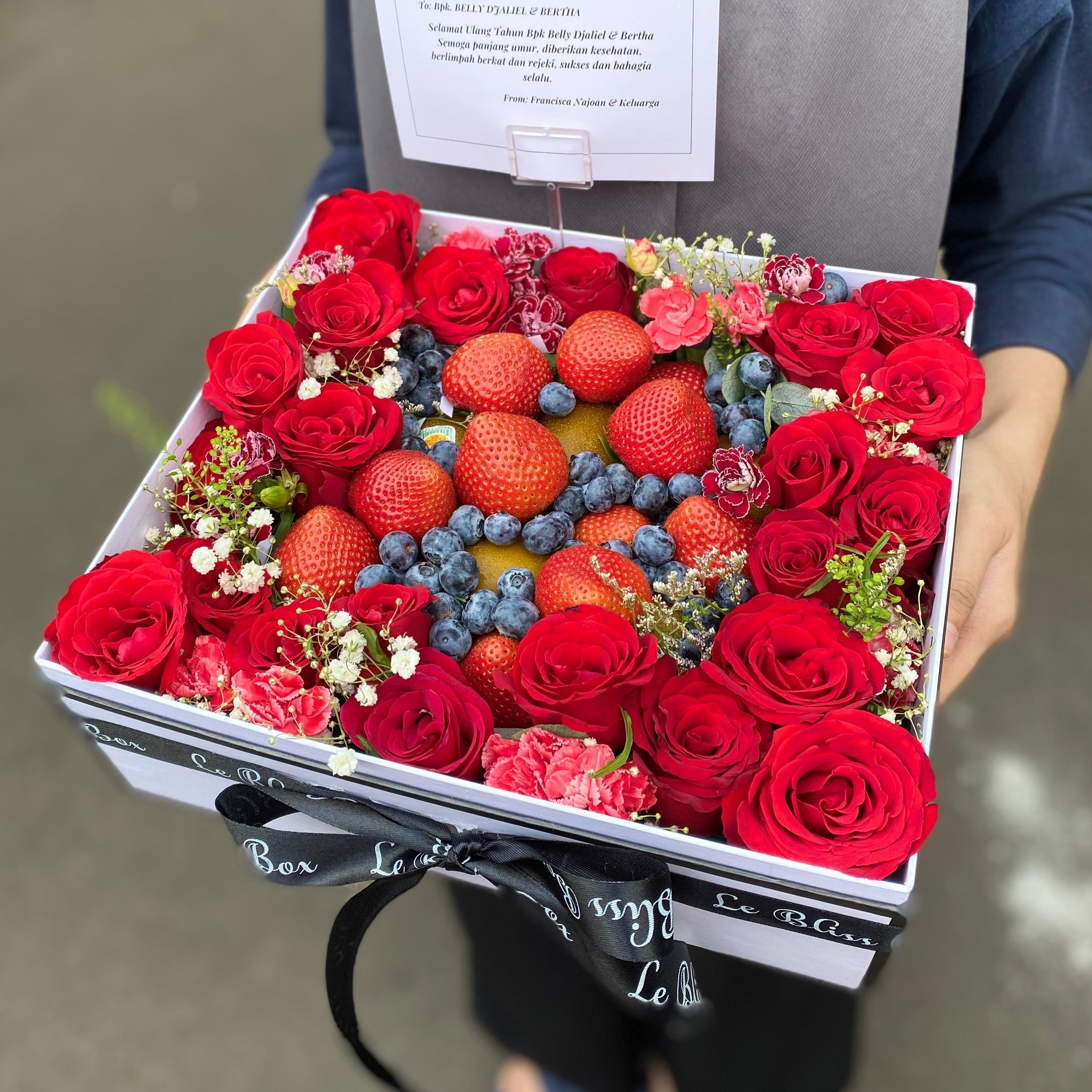 A flower box arrangement featuring an assortment of fresh berries and flowers, predominantly red and blue flowers, presented in a rectangular box with a black ribbon. Strawberry Blueberry Kiwi Flower Box – Parcel buah dan bunga segar elegan dari Le Bliss Bouquet Surabaya, cocok untuk hadiah ulang tahun dan hampers sehat. Parcel bUAH SURABAYA