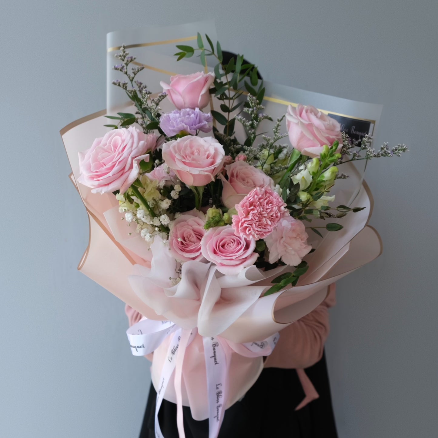 Bouquet of pink roses and other flowers with a white ribbon on a gray background. Le Bliss Bouquet’s Sweet Pink Medium Bouquet — elegant roses & carnations in soft pink tones. Same-day delivery across Jabodetabek, Surabaya & Cirebon.