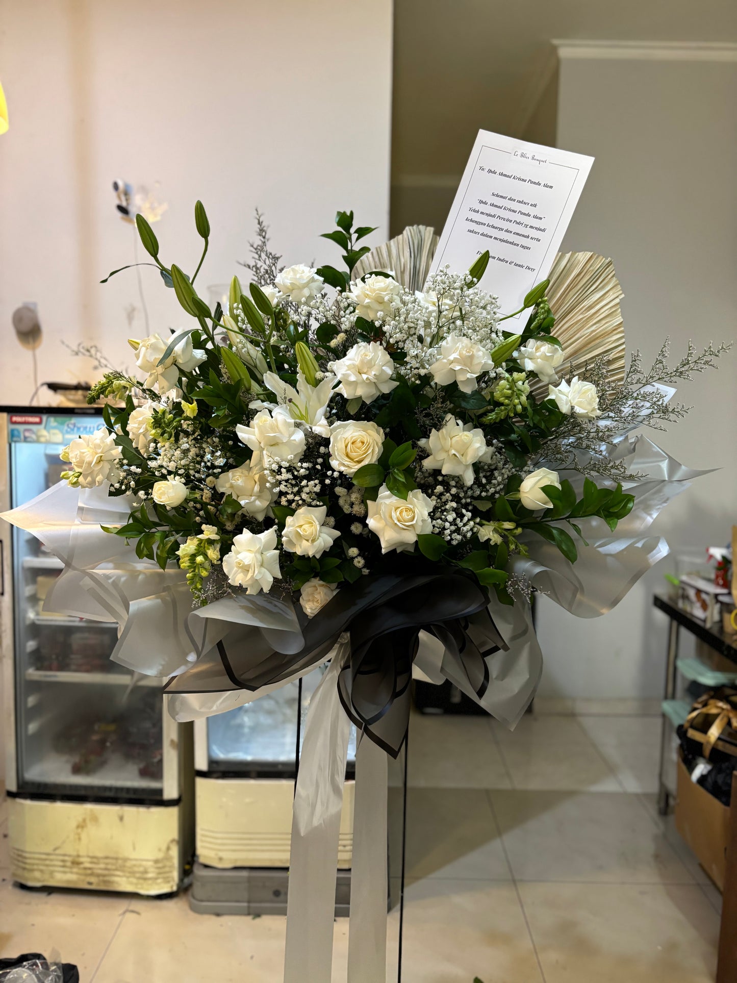 Bouquet of white flowers with greenery in a clear plastic wrap, placed indoors. Bouquet standing flower of white flowers with greenery on a stand in a store setting. florist surabaya, standing flower jakarta, standing flower surabaya