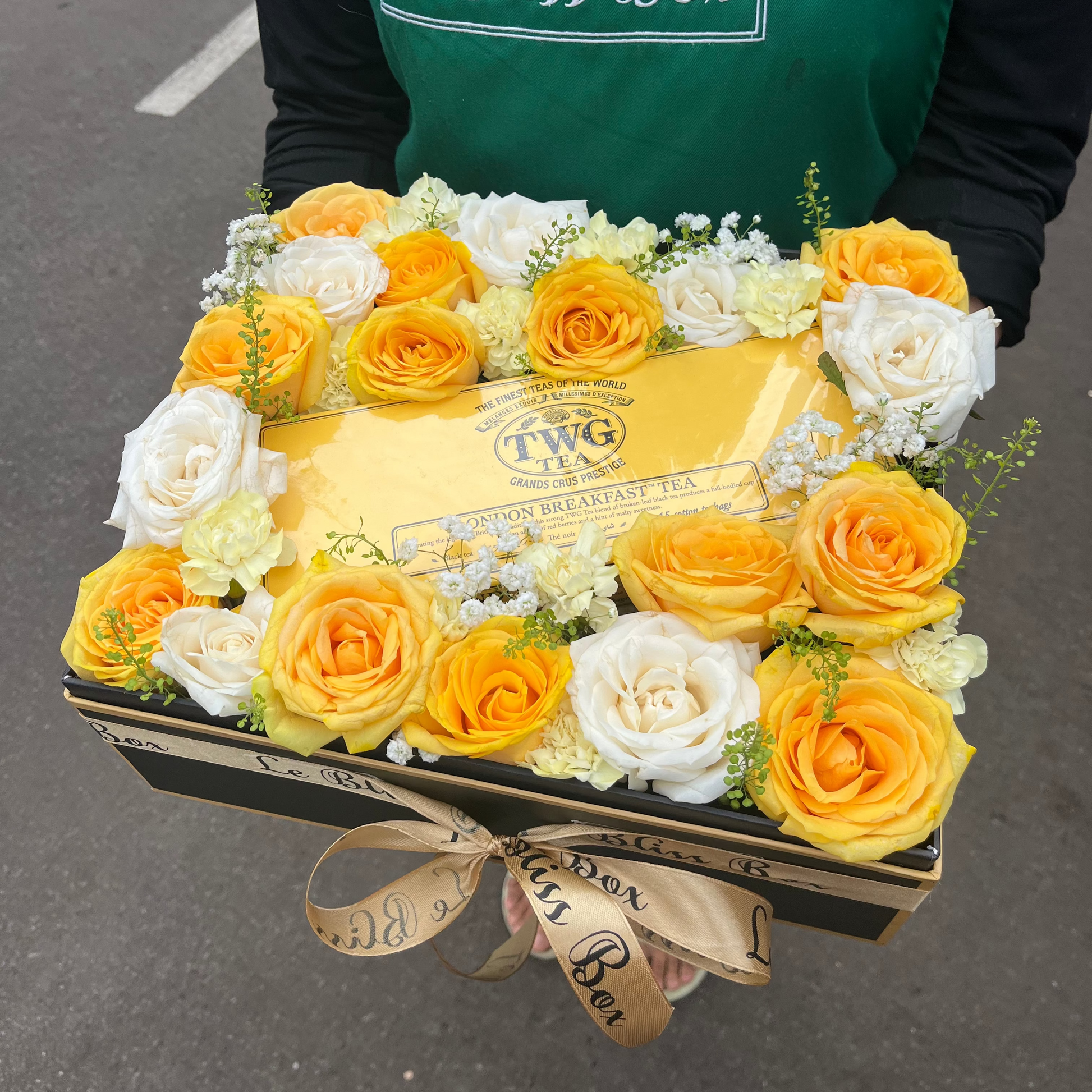 Box of TWG tea with yellow and white roses, held by a person wearing a green apron with 'Le Bliss Box' branding. TWG Hamper jakarta