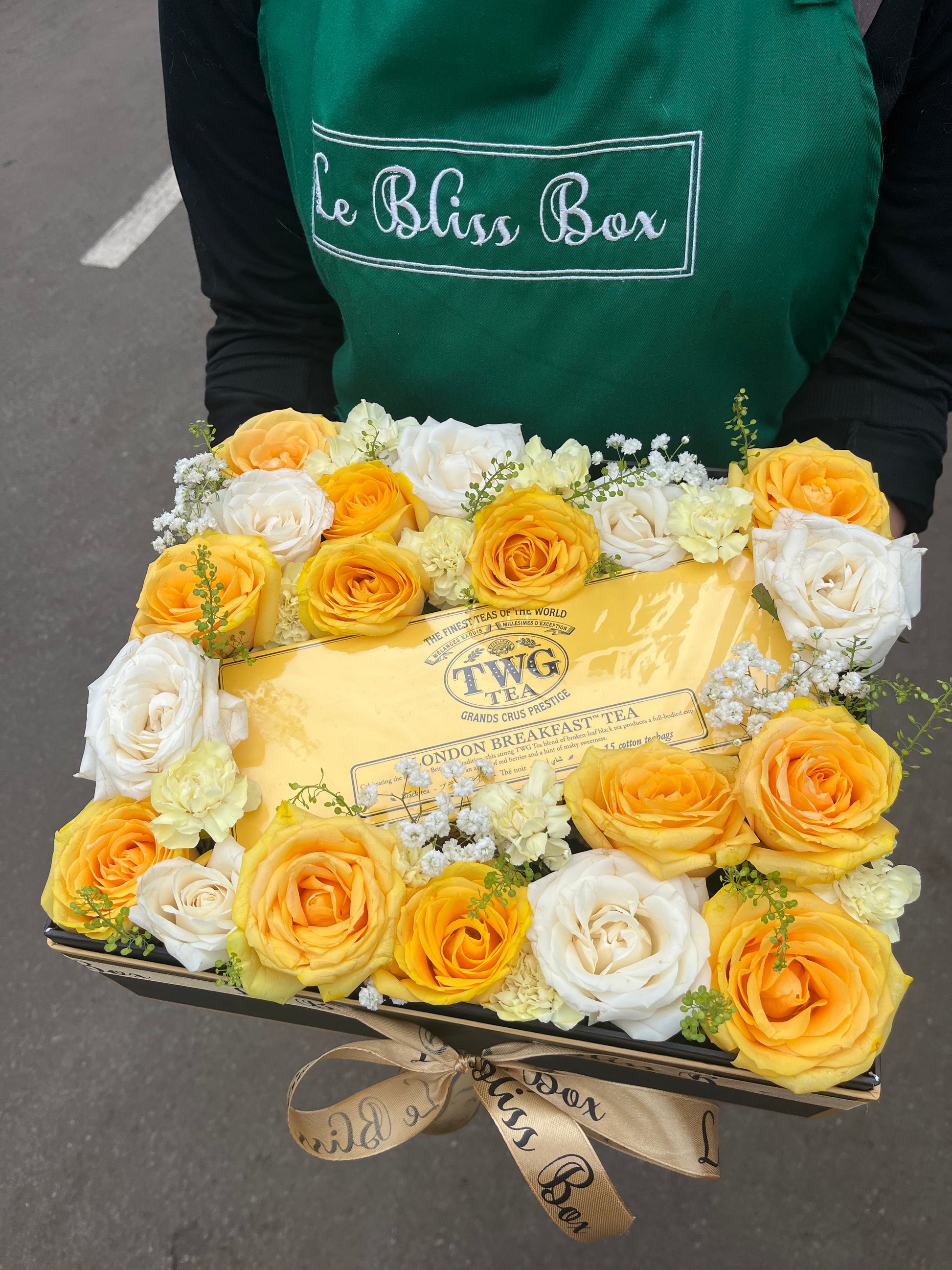 Box of TWG tea with yellow and white roses, held by a person wearing a green apron with 'Le Bliss Box' branding. twg hamper jakarta, surabaya