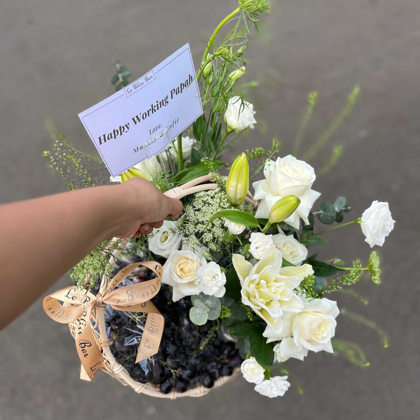 Keranjang rotan elegan berisi anggur segar, bunga lili, dan mawar putih yang indah, disusun dengan rapi untuk dijadikan hamper.