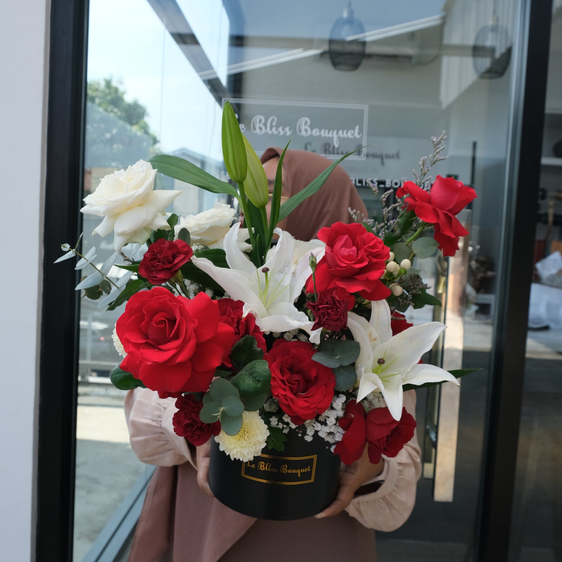 A person holding a round box containing an arrangement of red and white roses and lilies, with green leaves and decorative elements. florist surabaya, toko bunga surabaya, bloombox by le bliss bouquet