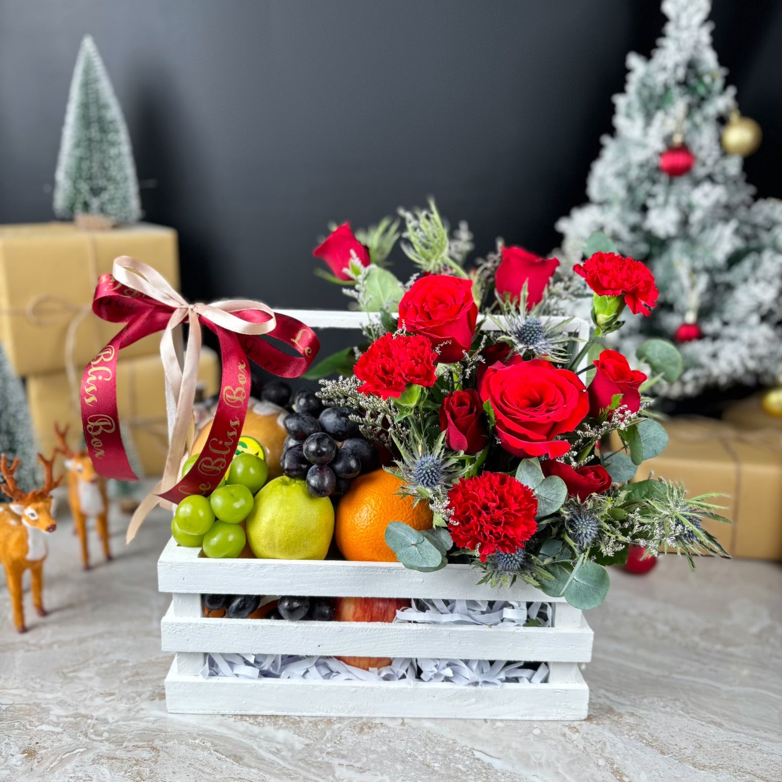 Decorative fruit and flower arrangement in a white crate with Christmas decorations in the background. Blooming Red Crate Fruit Parcel | Best Christmas Hamper in Jakarta & Surabaya – Le Bliss Box