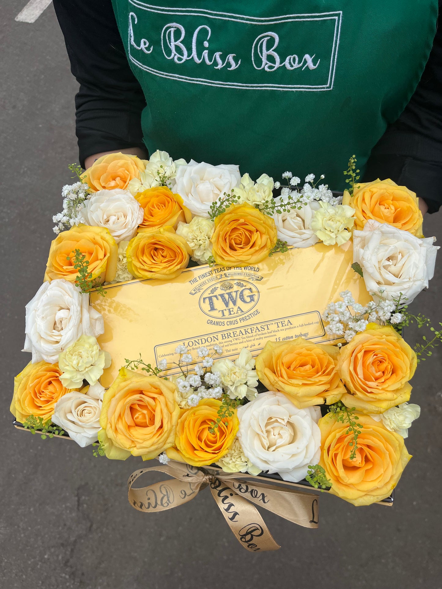 Floral arrangement with TWG tea box held by a person wearing a 'Le Bliss Box' apron. TWG Hamper le bliss box