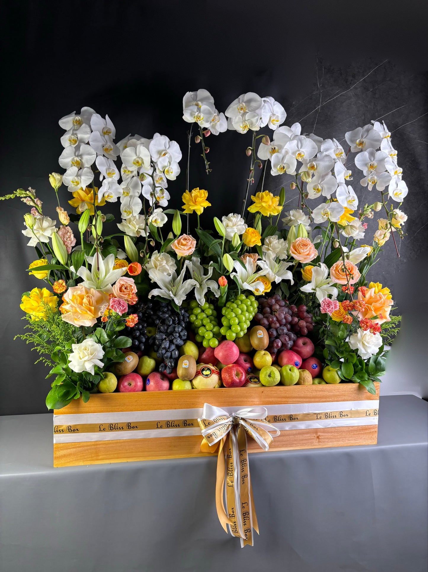 Floral arrangement with fruits in a decorative box on a dark background. Floral arrangement with fruits on a table, person adjusting flowers. Floral arrangement with fruits on a wooden stand against a dark background. rangkaian bunga dan parcel buah besar jakarta. by le bliss bouquet
