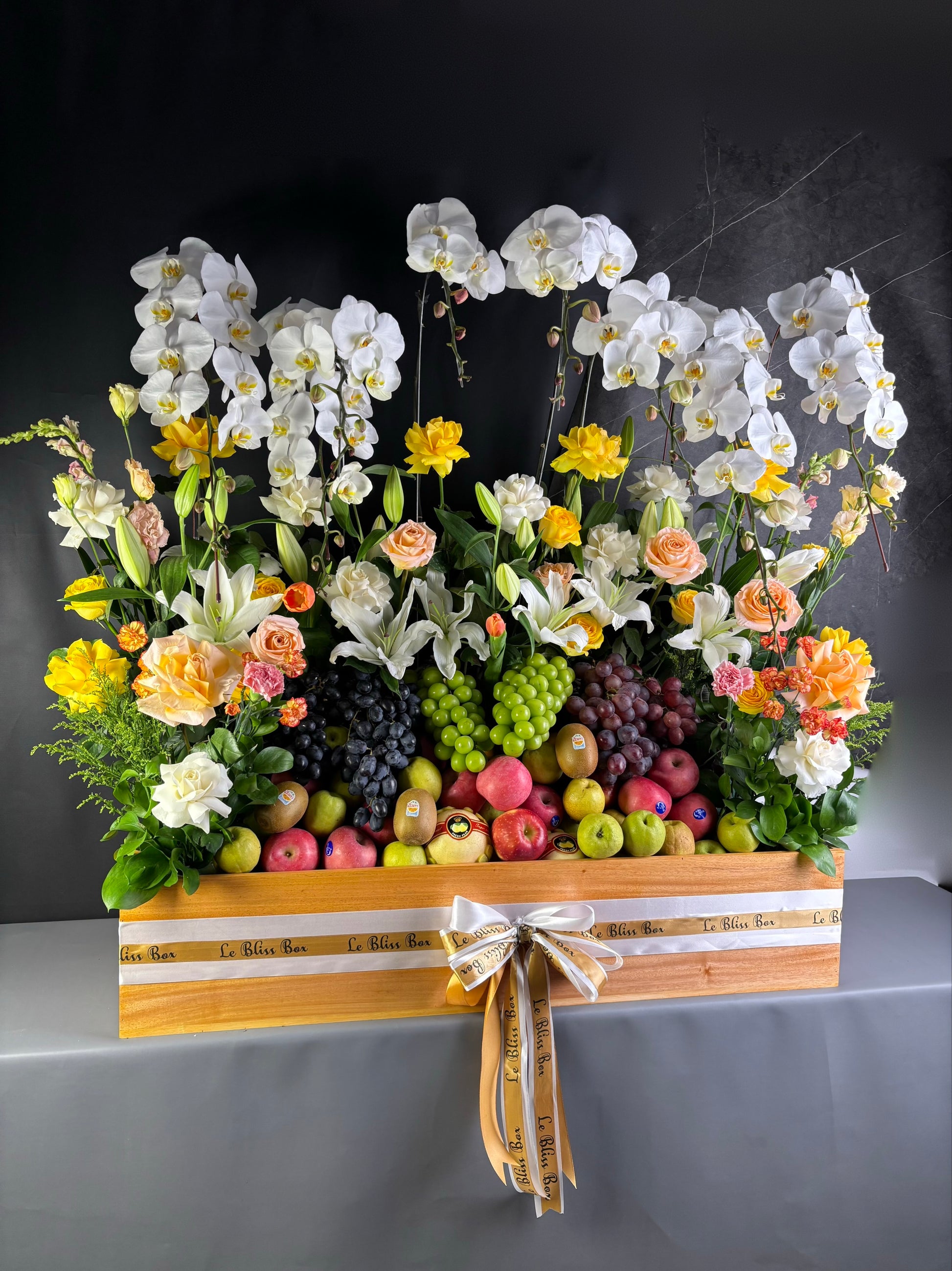 Floral arrangement with fruits in a decorative box on a dark background. Floral arrangement with fruits on a table, person adjusting flowers. Floral arrangement with fruits on a wooden stand against a dark background. rangkaian bunga dan parcel buah besar jakarta. by le bliss bouquet