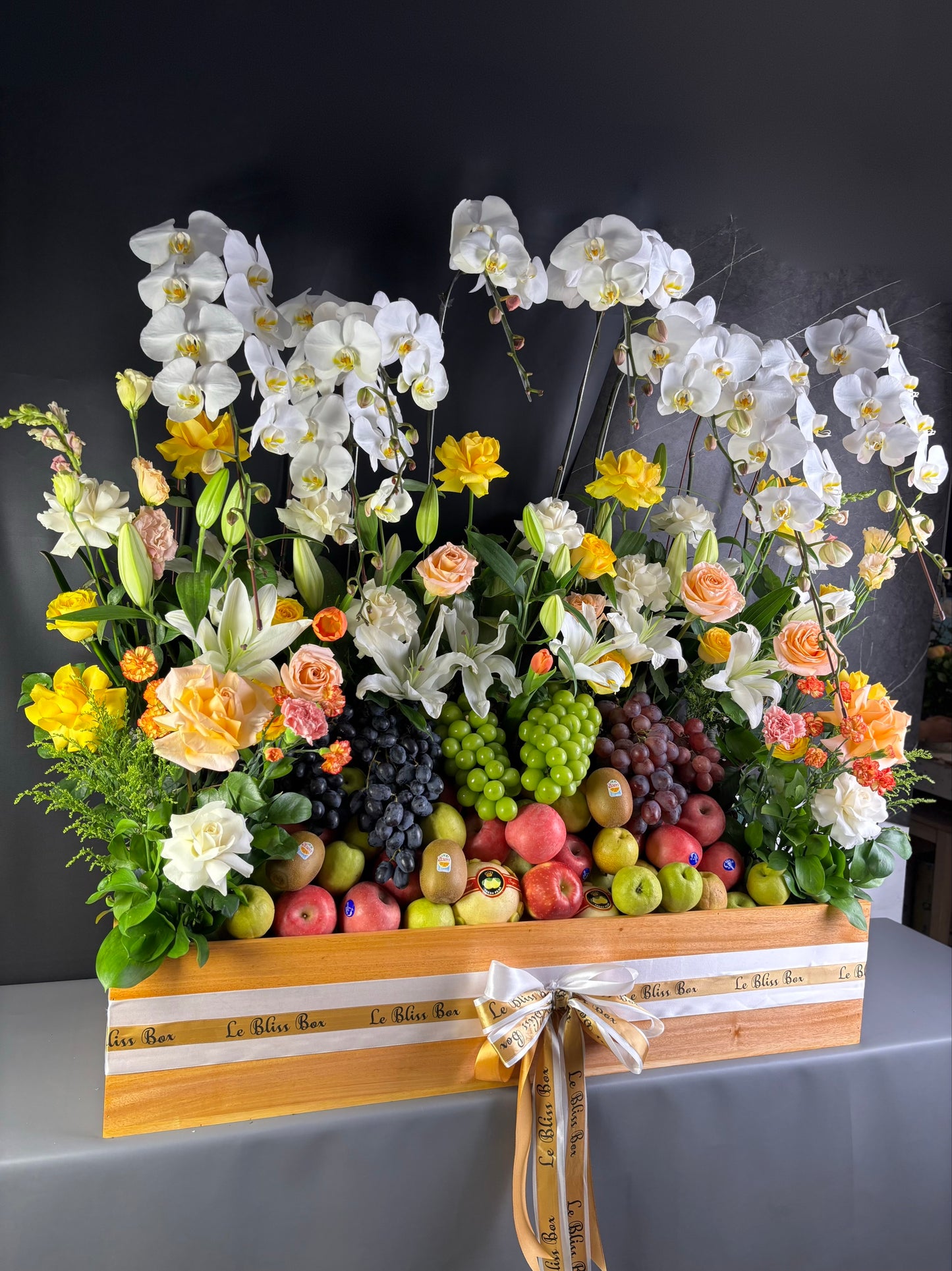 Floral arrangement with fruits in a wooden box on a dark background. Floral arrangement with fruits in a decorative box on a dark background. Floral arrangement with fruits on a table, person adjusting flowers. Floral arrangement with fruits on a wooden stand against a dark background. rangkaian bunga dan parcel buah besar jakarta. by le bliss bouquet