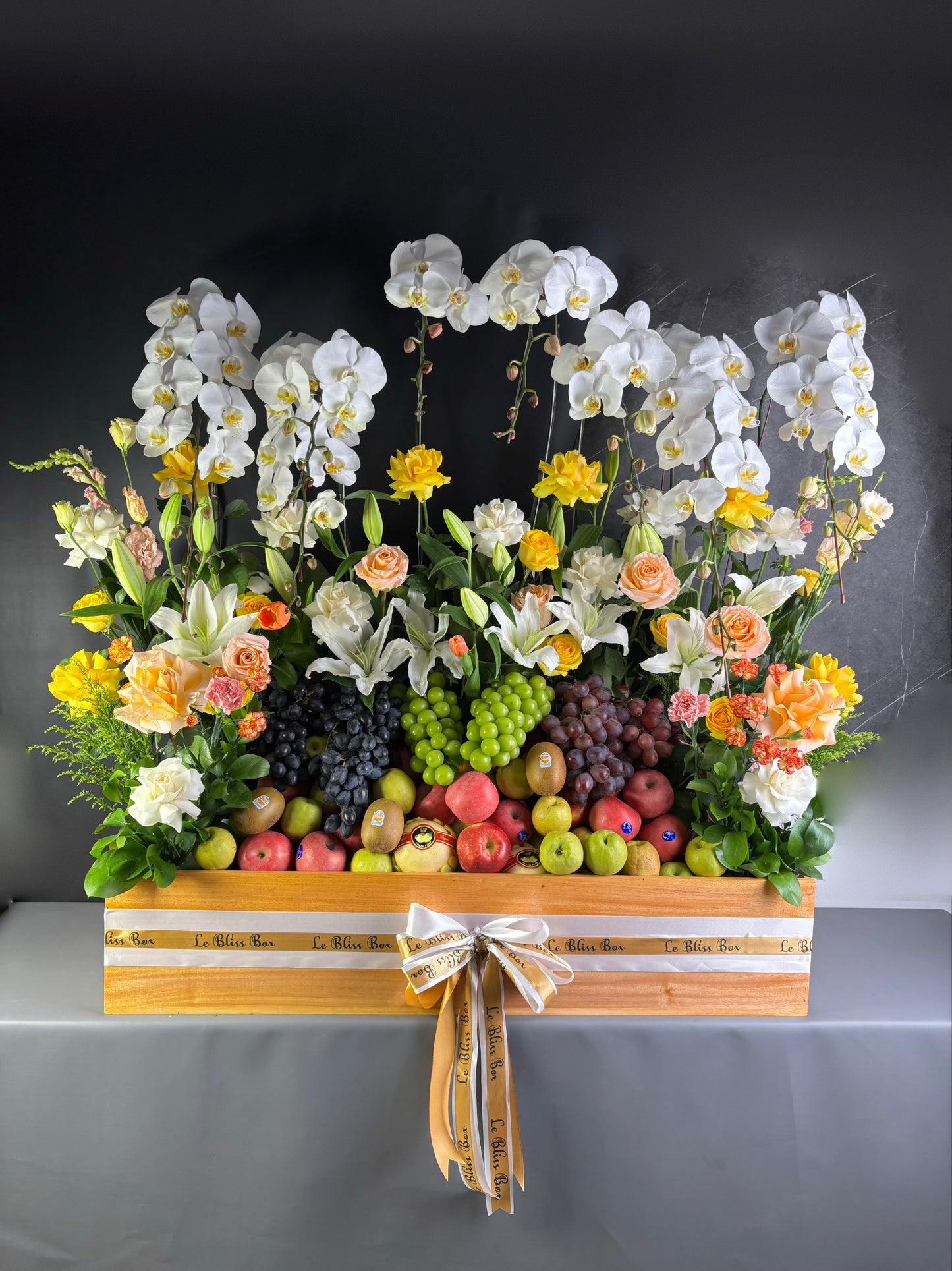 Floral arrangement with fruits on a wooden stand against a dark background. rangkaian bunga dan parcel buah besar jakarta. by le bliss bouquet