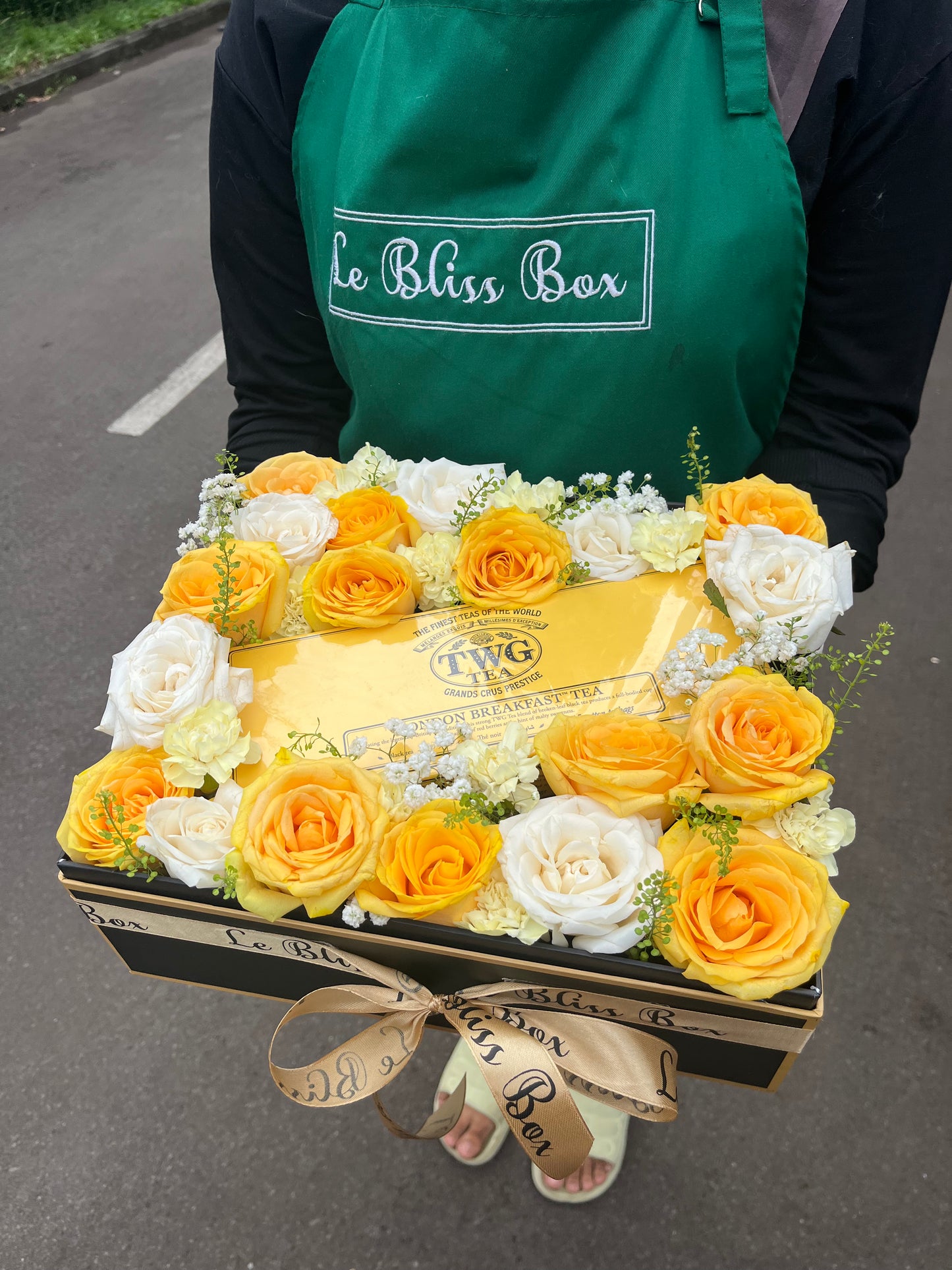 Floral arrangement with yellow and white roses and a TWG tea box, held by a person wearing a green apron with 'Le Bliss Box' branding. TWG Hamper mawar kuning putih jakarta surabaya