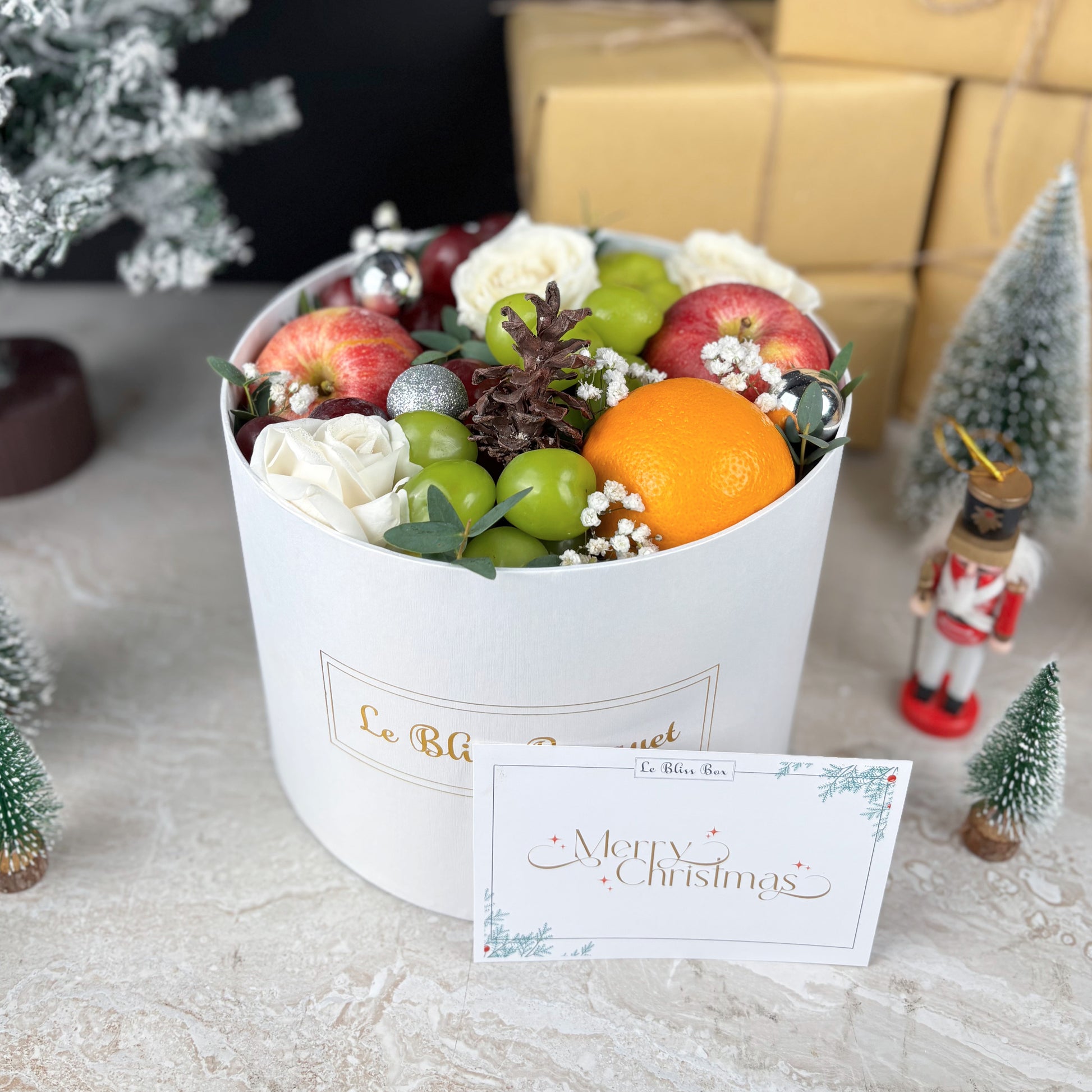 Fruit and flower arrangement in a white box with a 'Merry Christmas' card, surrounded by festive decorations. Winter Sonata Round Fruit Box dari Le Bliss Box. Christmas fruit hamper elegan dengan buah premium. Pengiriman Surabaya, Jakarta & Cirebon. Luxury, festive, and fresh.