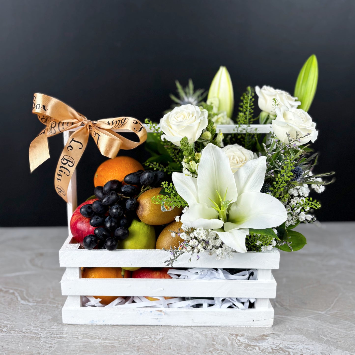 Fruit and flower arrangement in a white crate with a gold ribbon on a dark background. Blooming White Crate Fruit Parcel | Best Christmas Hamper Jakarta, Tangerang, Cirebon & Surabaya – Le Bliss Bouquet