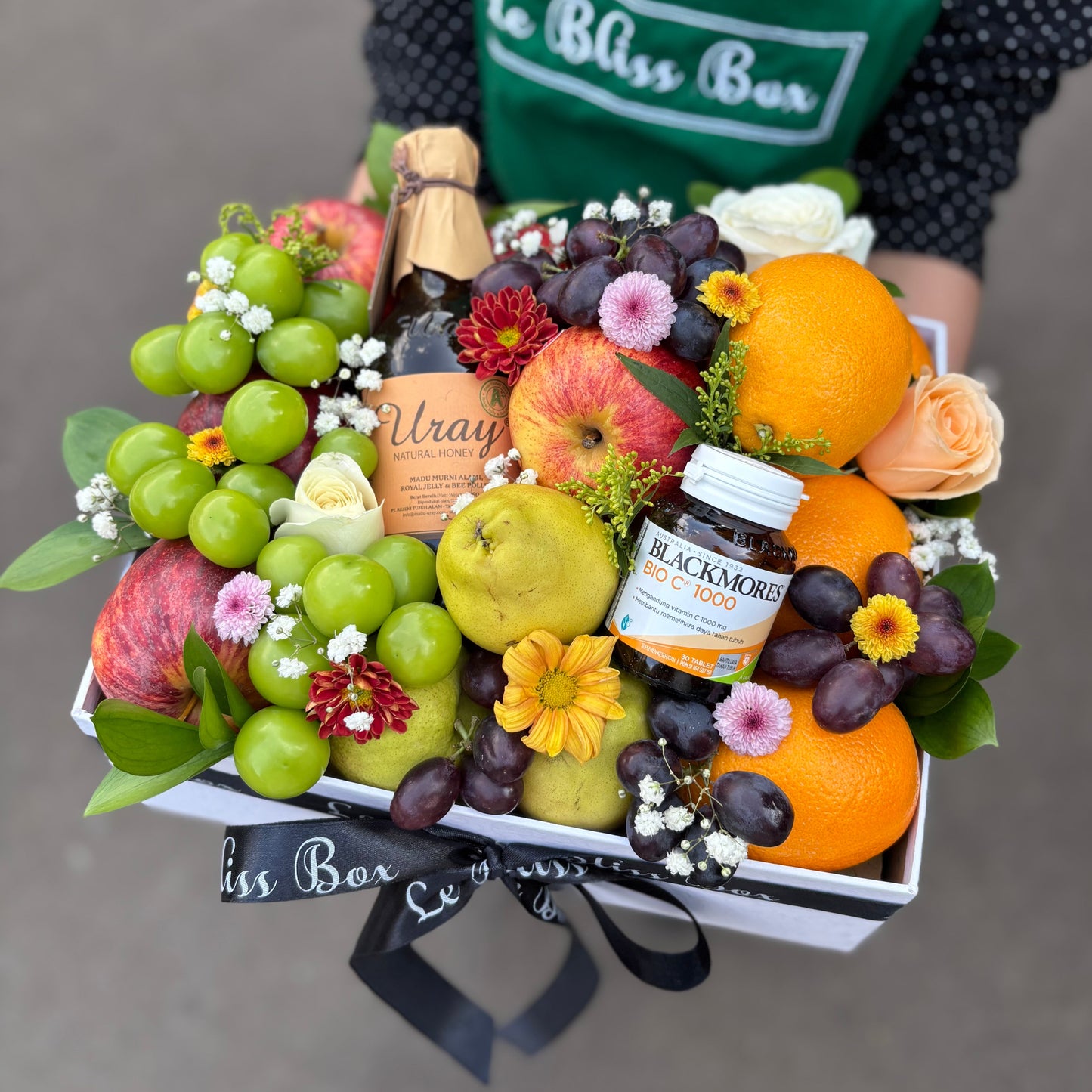 Fruit and flower arrangement with a bottle of Blackmores supplement, held by a person wearing a green apron with 'Le Bliss Box' branding. black mores vitamin c with honey fruit hamper surabaya, Jakarta, sidoarjo. parcel buah surabaya, jakarta, bintaro
