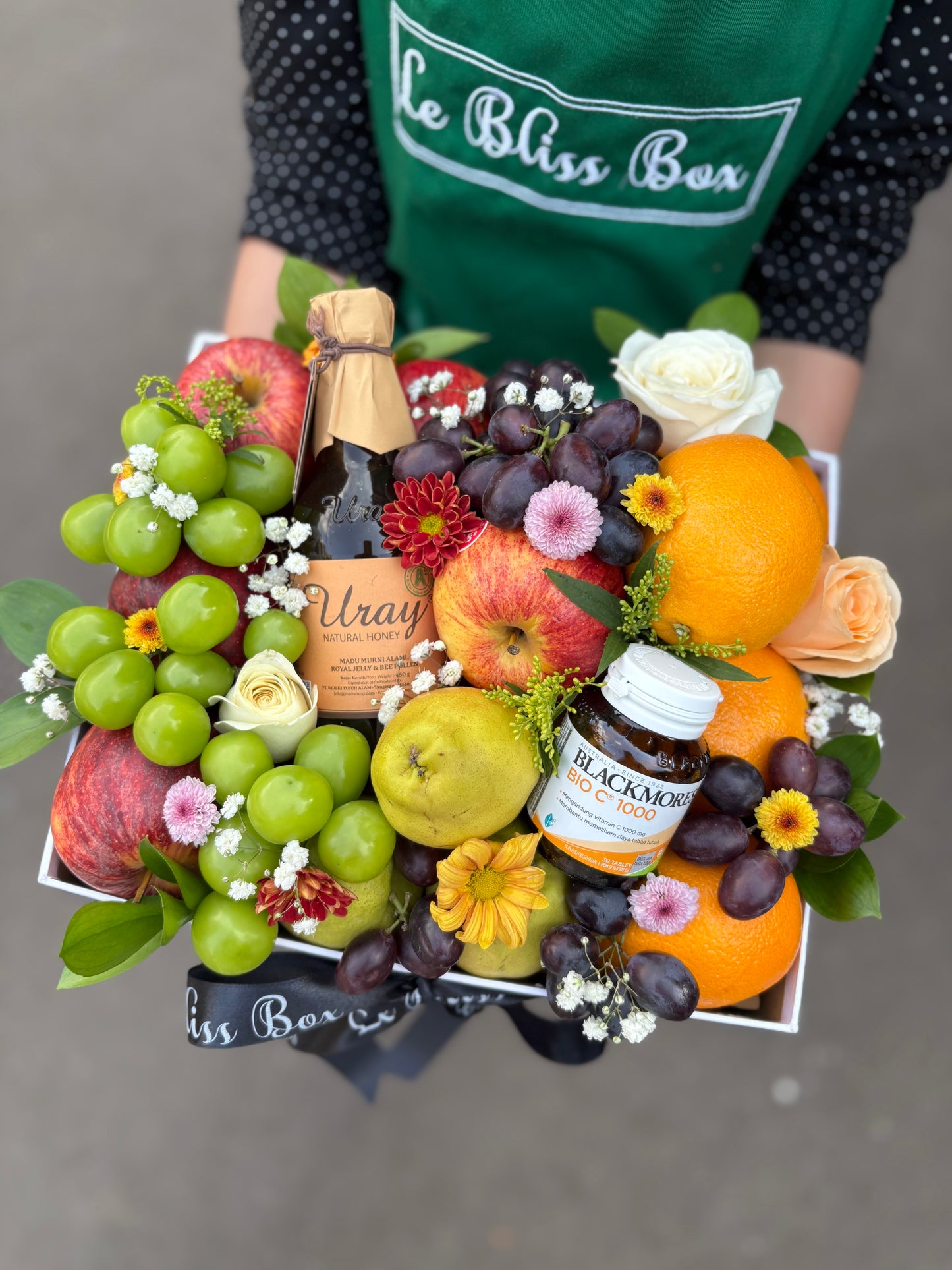 Fruit and flower arrangement with a person wearing a 'Le Bliss Box' apron. Person holding a box of fruits, flowers, and a bottle of wine with 'Le Bliss Box' branding. Parcel buah surabaya, parcel buah jakarta, parcel buah blackmores uray bintaro