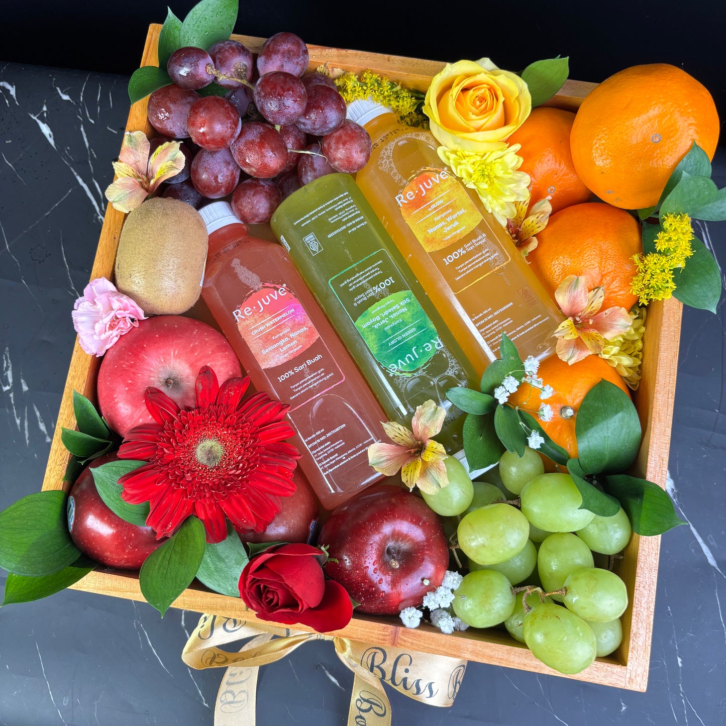 A wooden fruit box containing an assortment of fresh fruits such as apples, oranges, grapes, and bananas, along with two bottles of juice and decorative flowers. rejuve juice fruit hamper jakarta, surabaya, cirebon