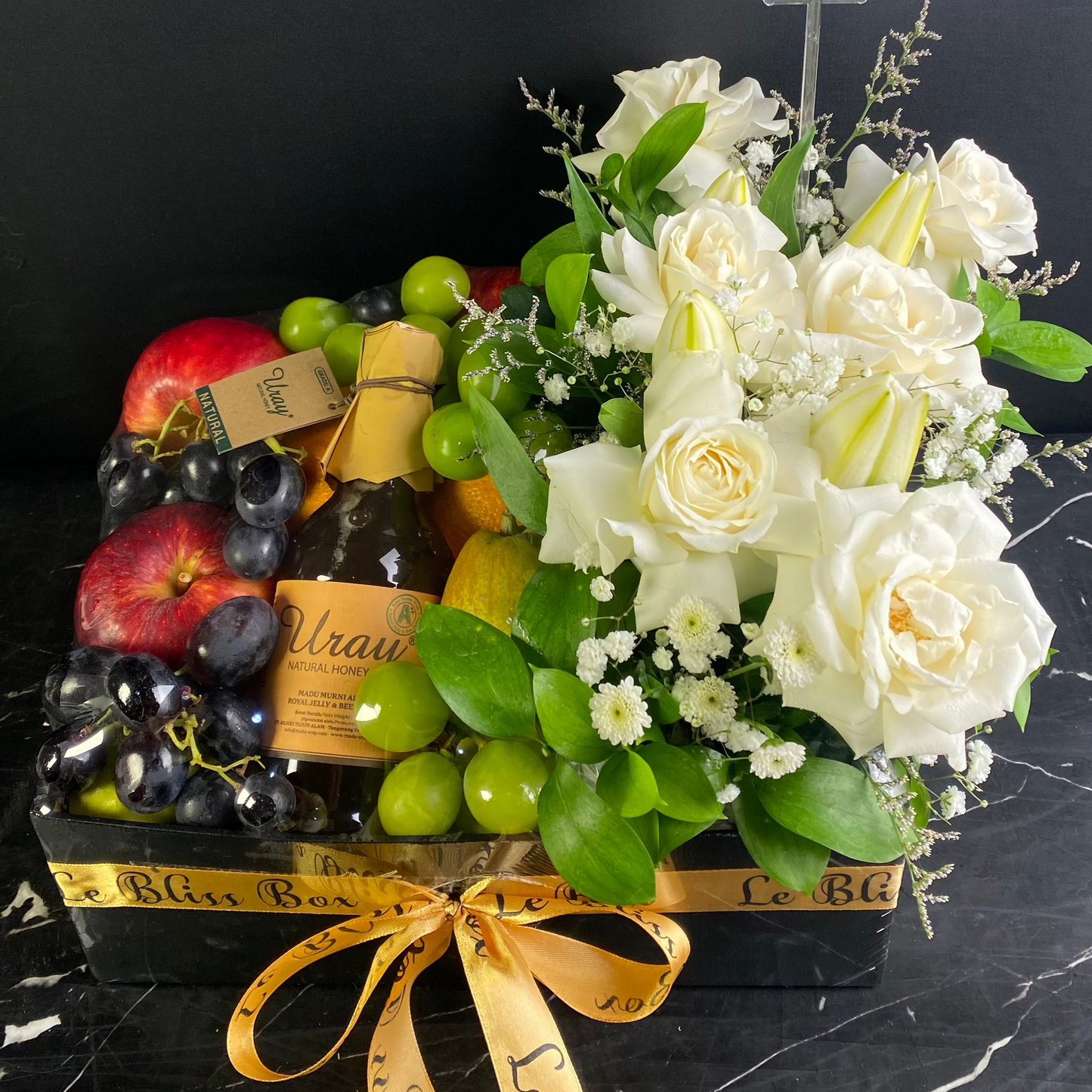 Gift basket with flowers, fruits, and a bottle of wine on a black surface. le Bliss box, honey fruit box. parcel buah madu surabaya