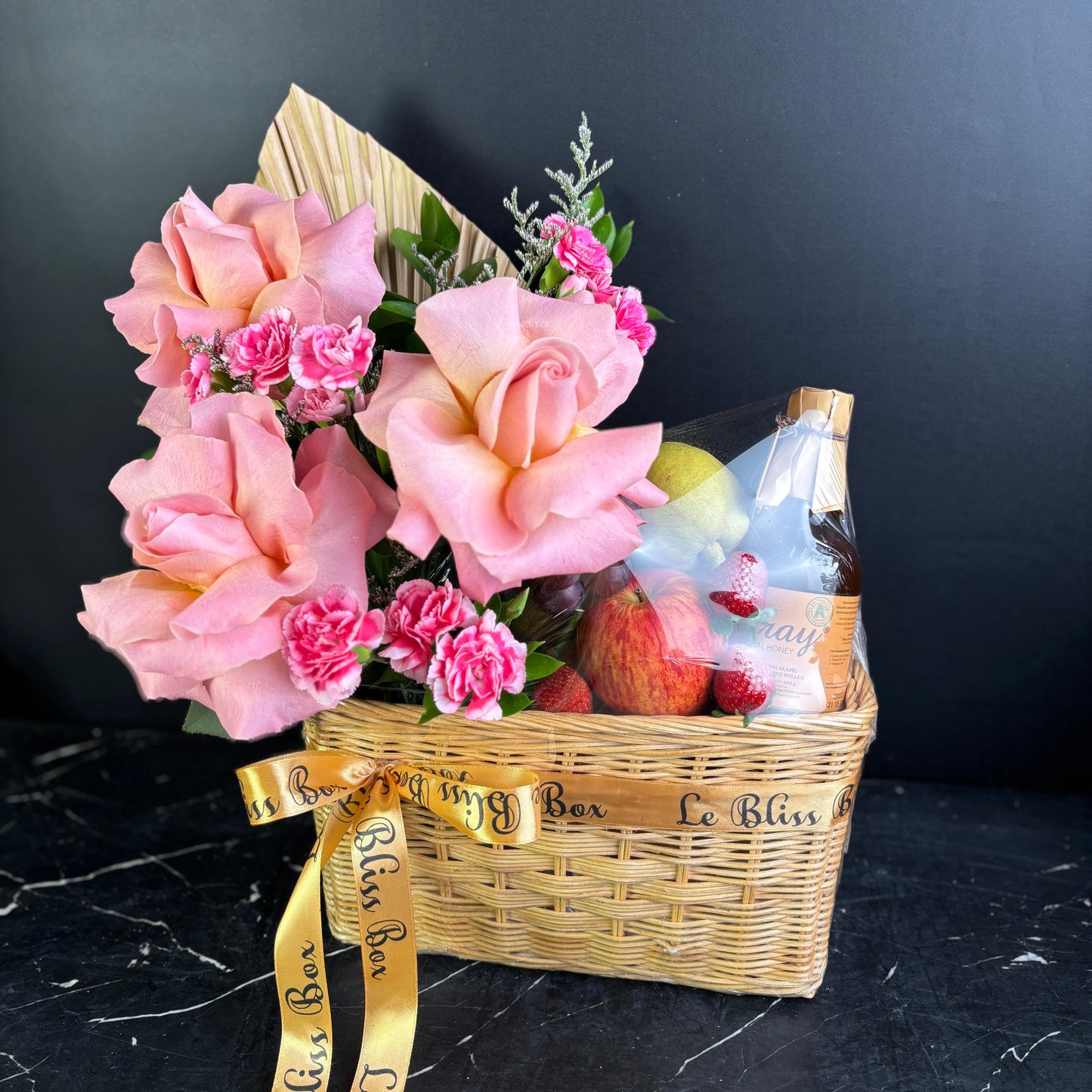 Gift basket with pink flowers, fruits, and a bottle on a dark surface. parcel buah surabaya dengan madu. parcel buah jakarta