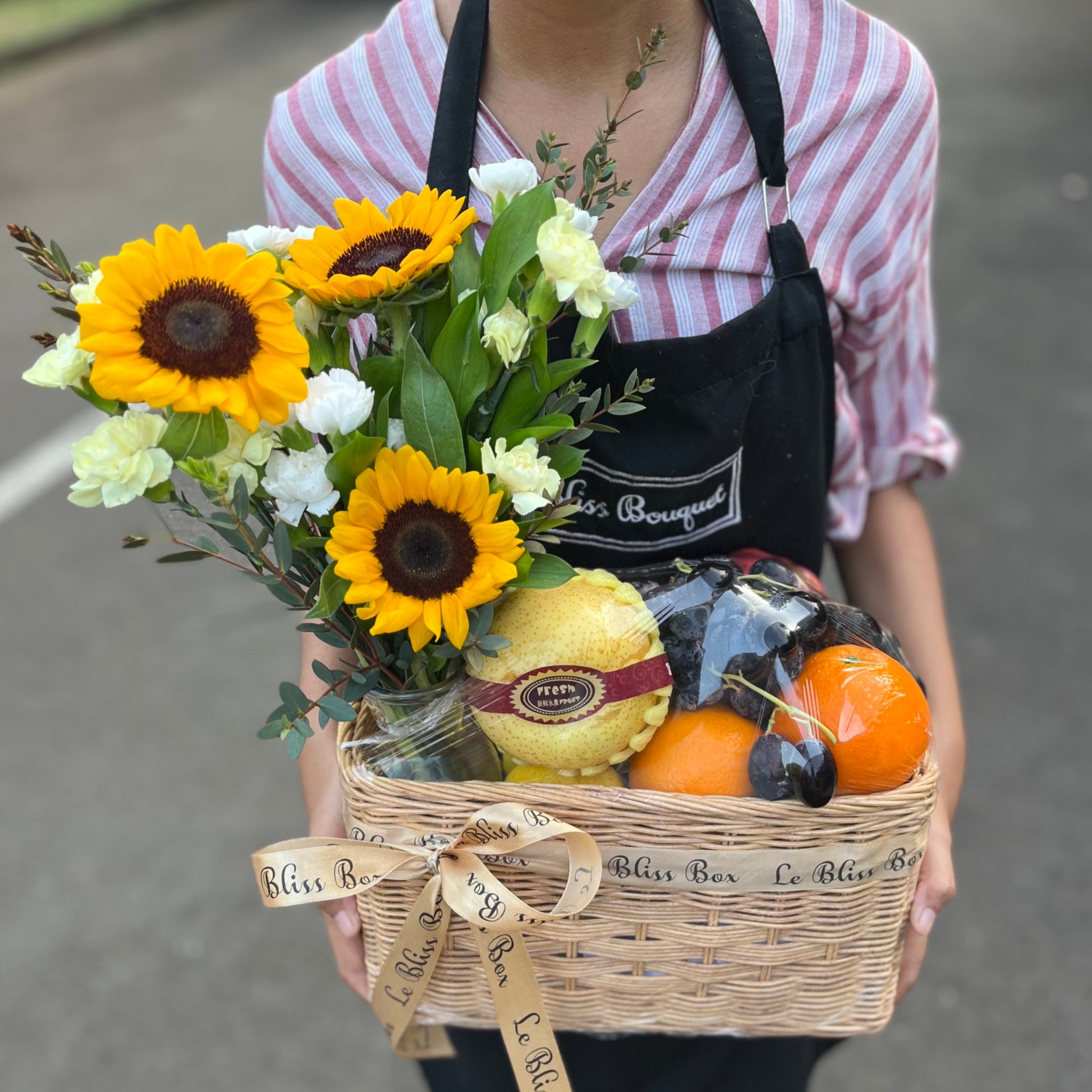 A person holding a basket with fresh sunflowers and various fruits, presented as a gift. Parcel buah surabaya, fruit hamper jabodetabek, surabaya