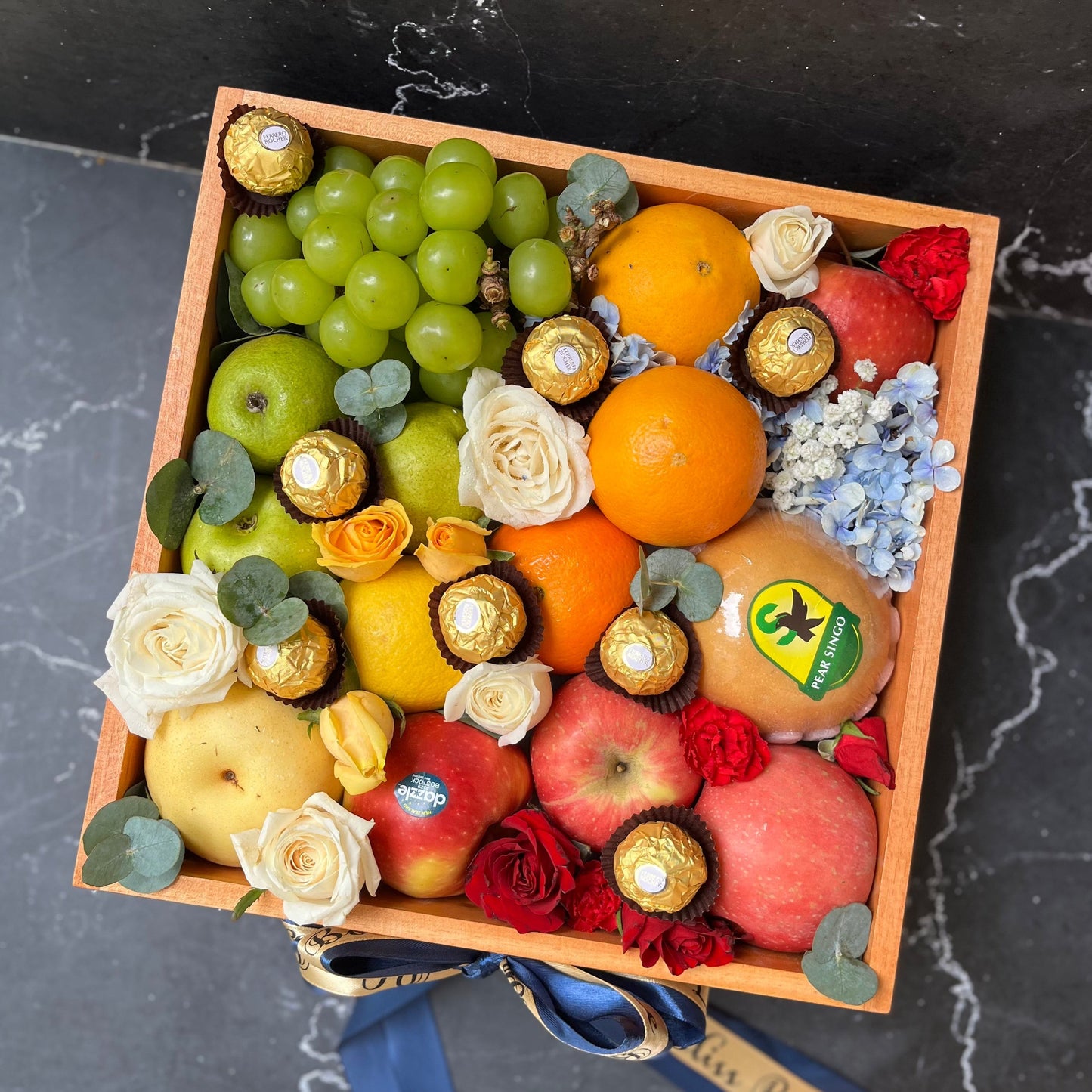 A wooden fruit and chocolate box arranged with an assortment of fruits including grapes, oranges, apples, and pears, and chocolate pieces, decorated with flowers and a bow with a tag. parcel buah surabaya