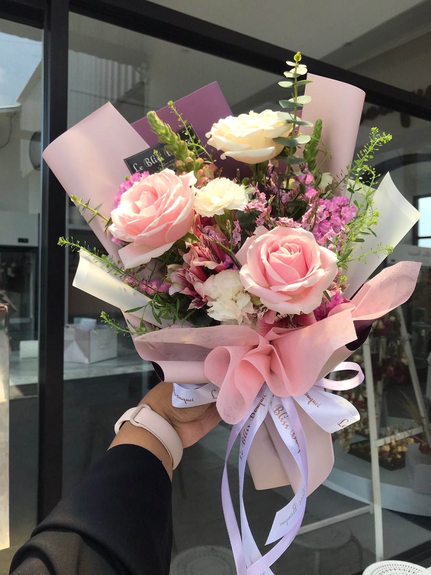 A small bouquet of flowers including white and pink roses, with a touch of lush purple, held in front of a store window. Le Bliss Bouquet Surabaya. toko bunga surabaya, florist surabaya, bouquet surabaya, Buket Bunga surabaya