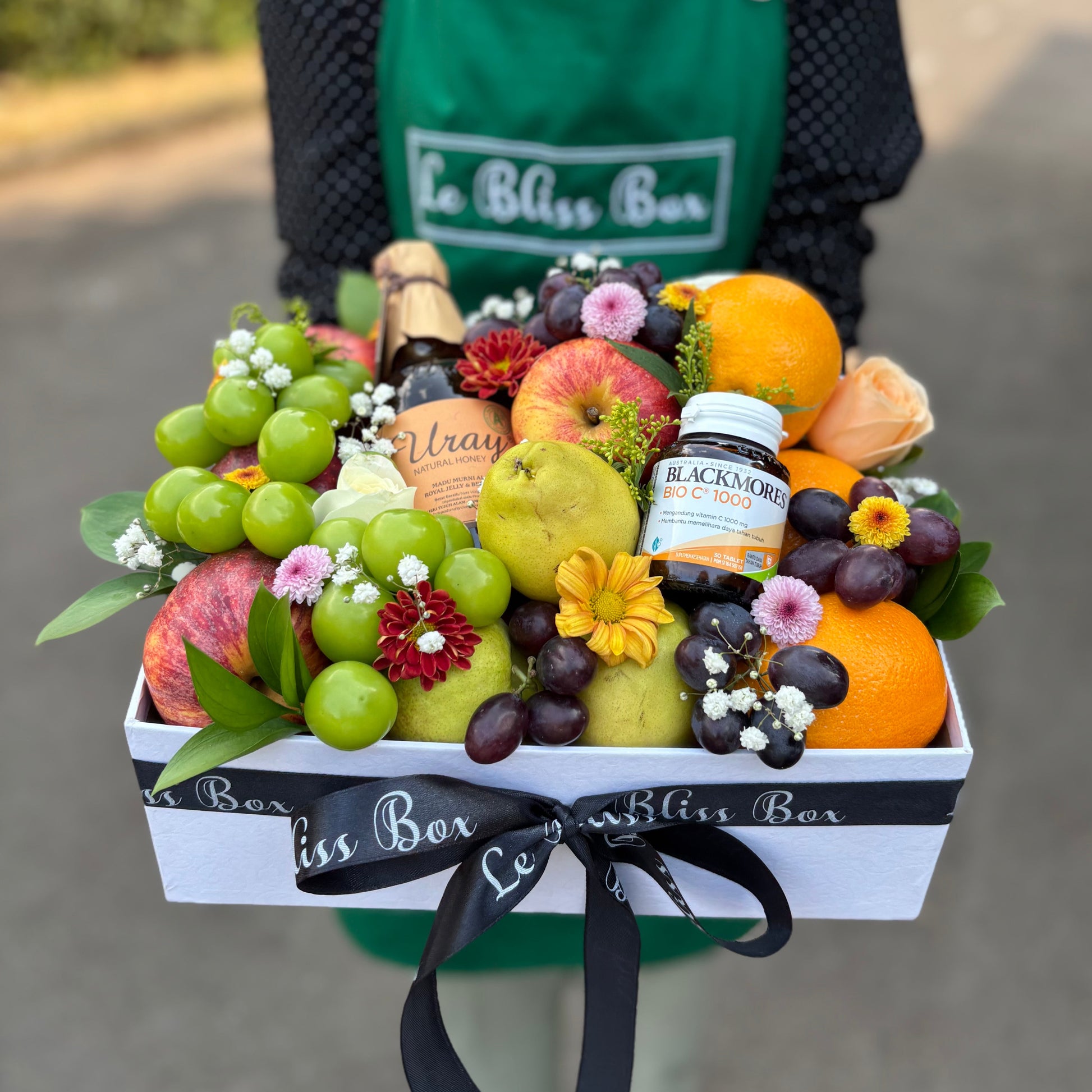 Person holding a box of fruits, flowers, and a bottle of wine with 'Le Bliss Box' branding. Parcel buah surabaya, parcel buah jakarta, parcel buah blackmores uray bintaro