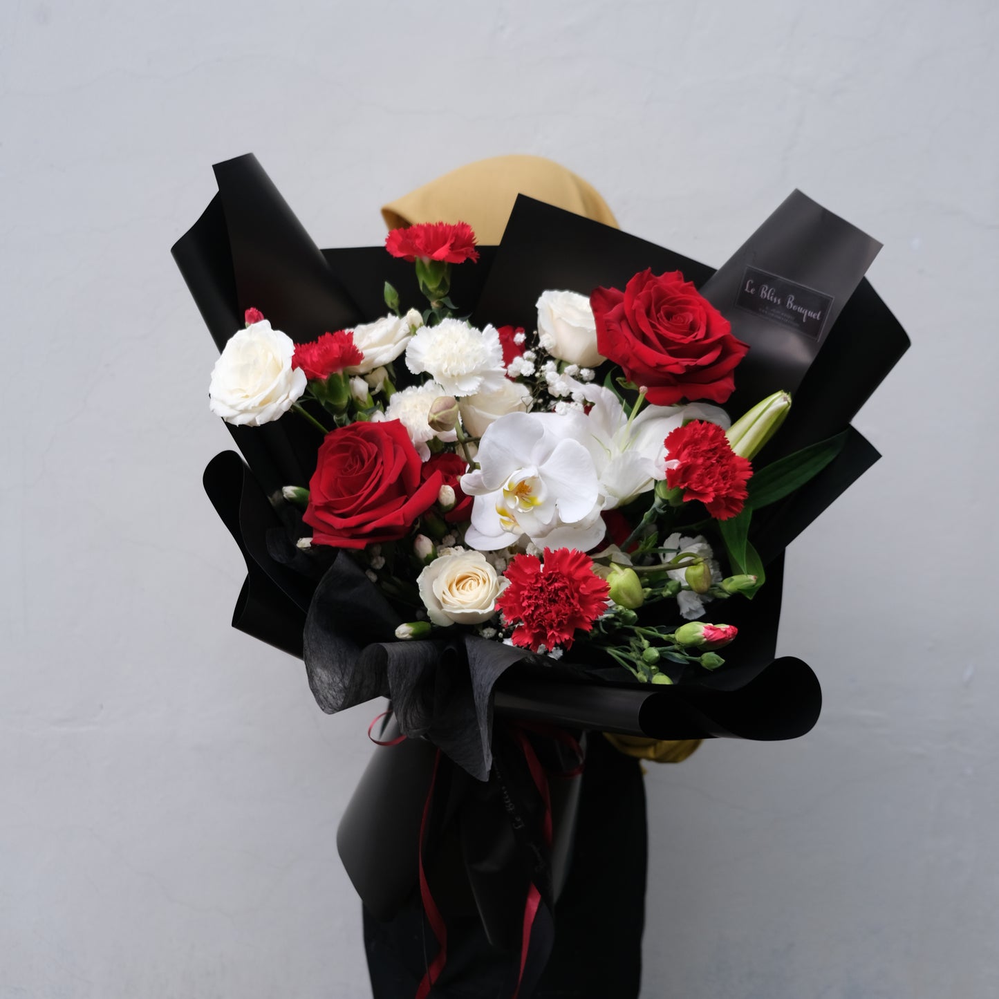 Rangkaian buket elegan dengan mawar merah dan putih serta anggrek putih, dibalut dengan kertas hitam dari Le Bliss Bouquet