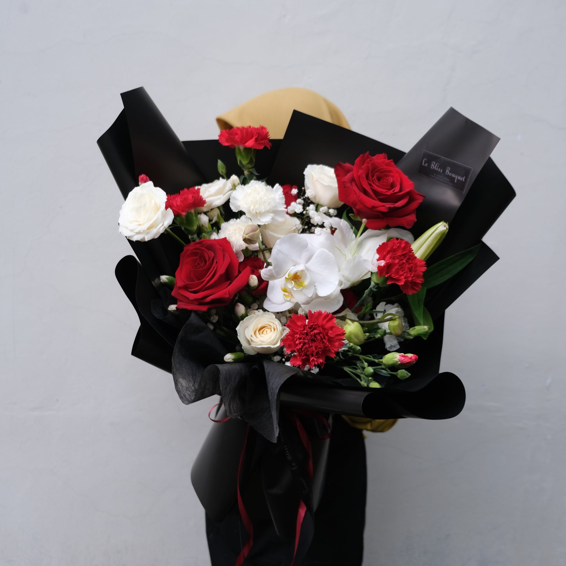 Rangkaian buket elegan dengan mawar merah dan putih serta anggrek putih, dibalut dengan kertas hitam dari Le Bliss Bouquet