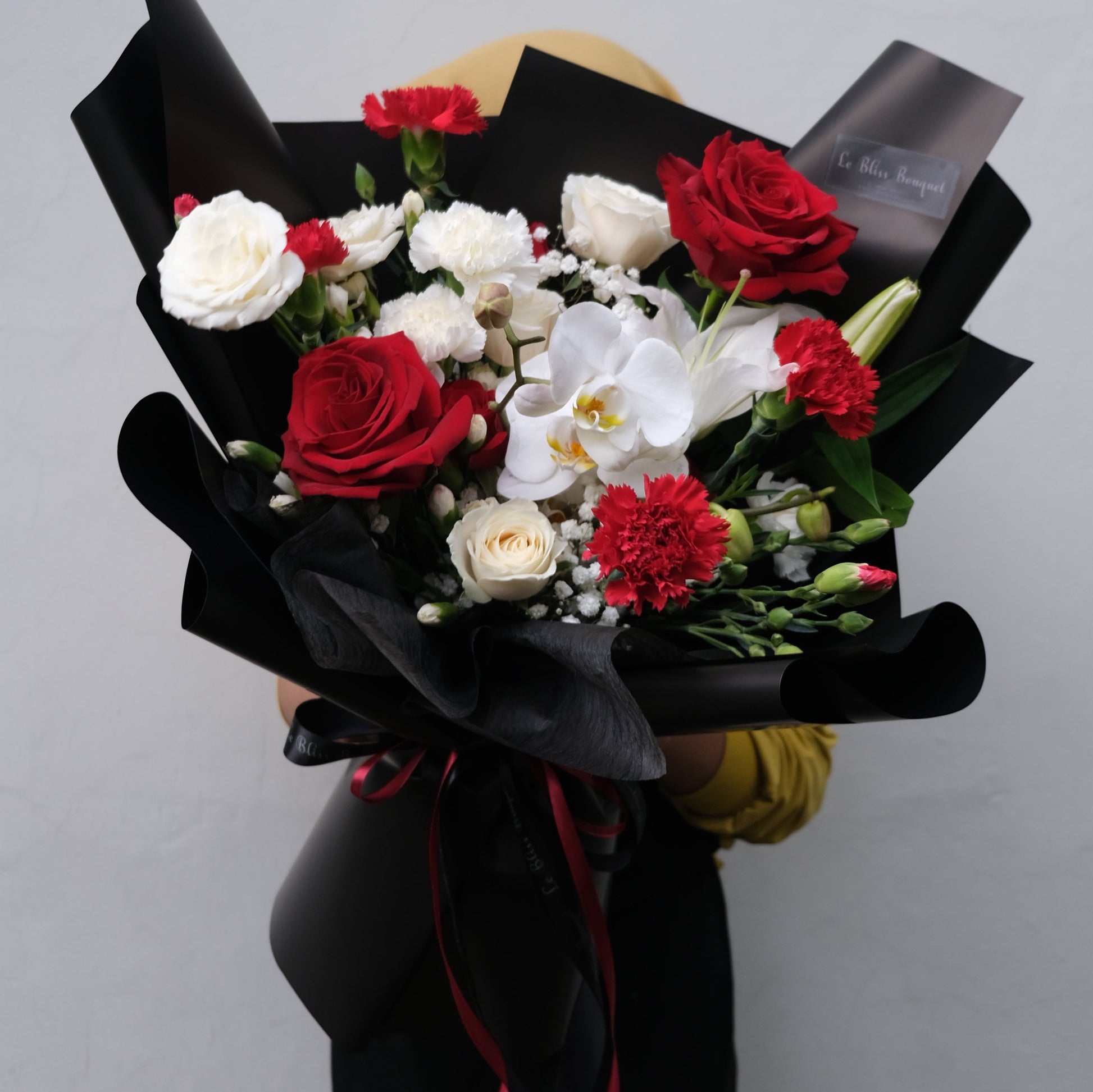Rangkaian buket elegan dengan mawar merah dan putih serta anggrek putih, dibungkus dengan kertas hitam dari Le Bliss Bouquet