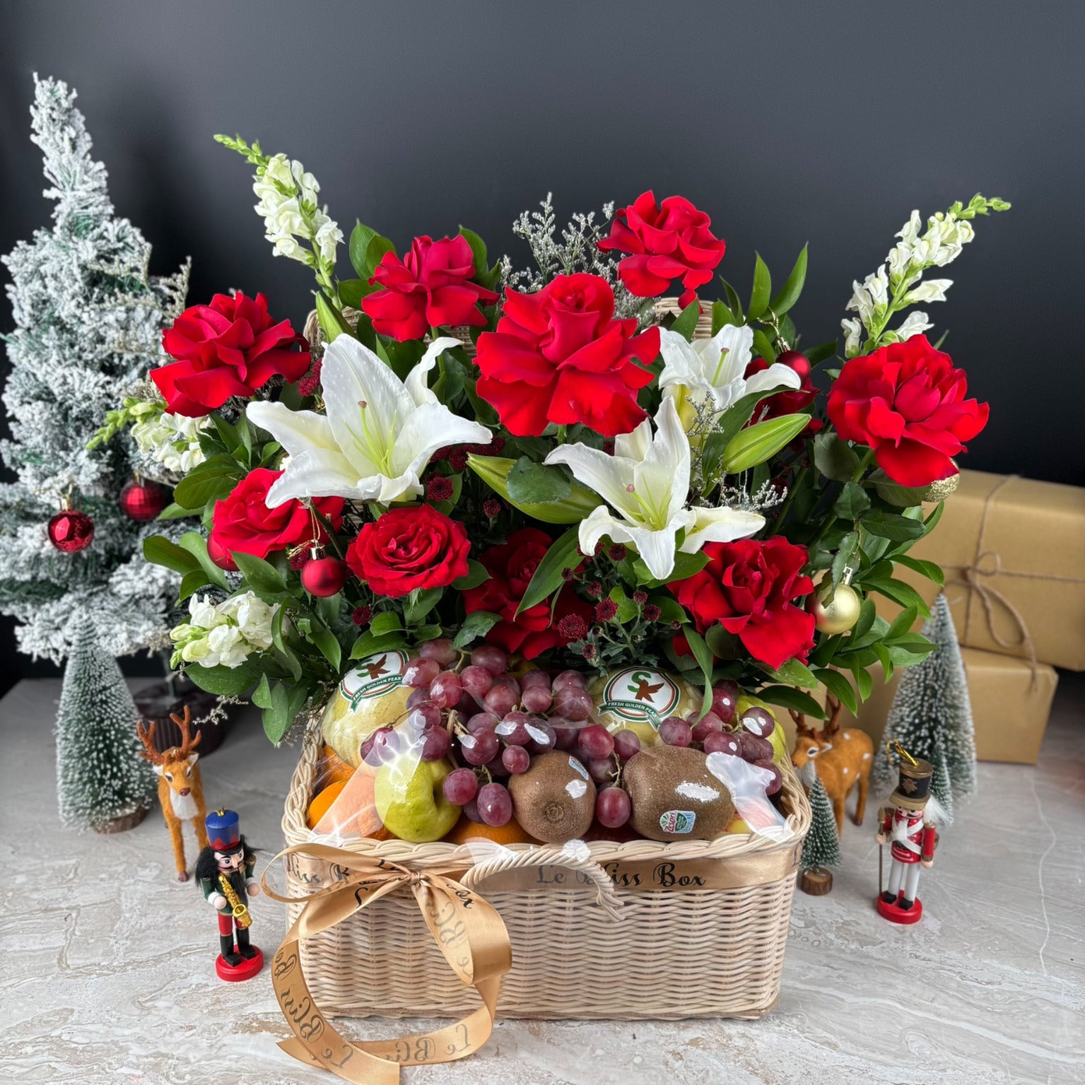 hamper of red and white flowers with a fruit basket on a decorative setting with Christmas trees and ornaments. Celebrate Christmas with Le Bliss Box’s Festive Grand Fruit Parcel — a luxurious fruit hamper for Jakarta, Surabaya & Tangerang delivery.