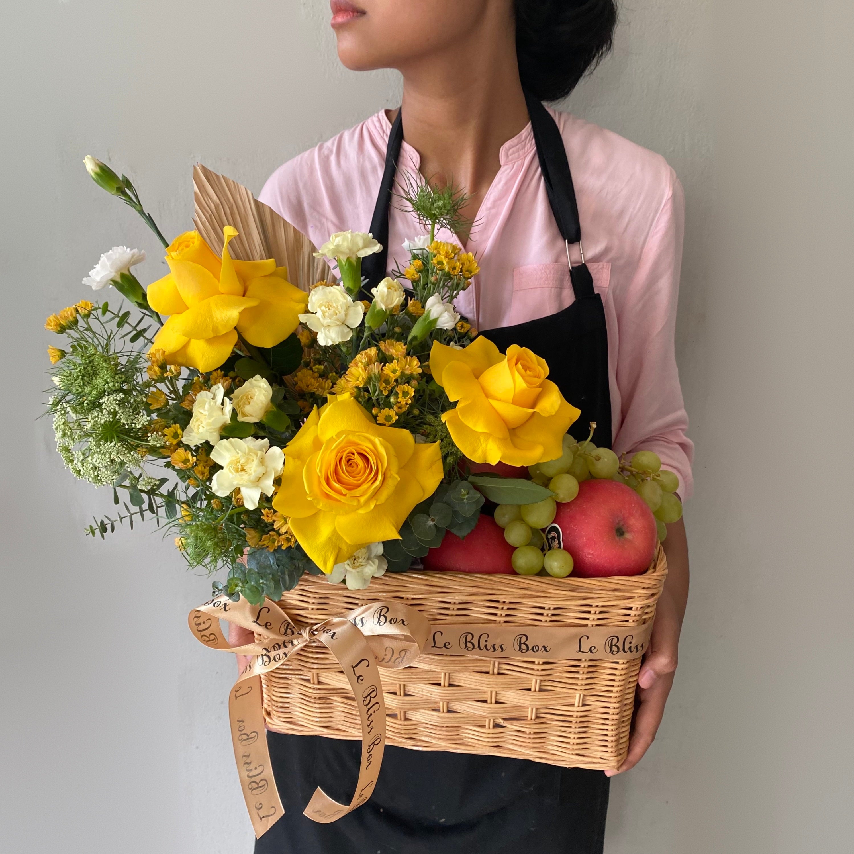 Keranjang anyaman cantik berisi buah-buahan segar dan bunga mawar kuning yang memikat, dipegang oleh seorang wanita dengan kemeja merah muda. Tersedia untuk pengiriman di Jakarta, Bintaro, Bogor, Bekasi, dan Depok.