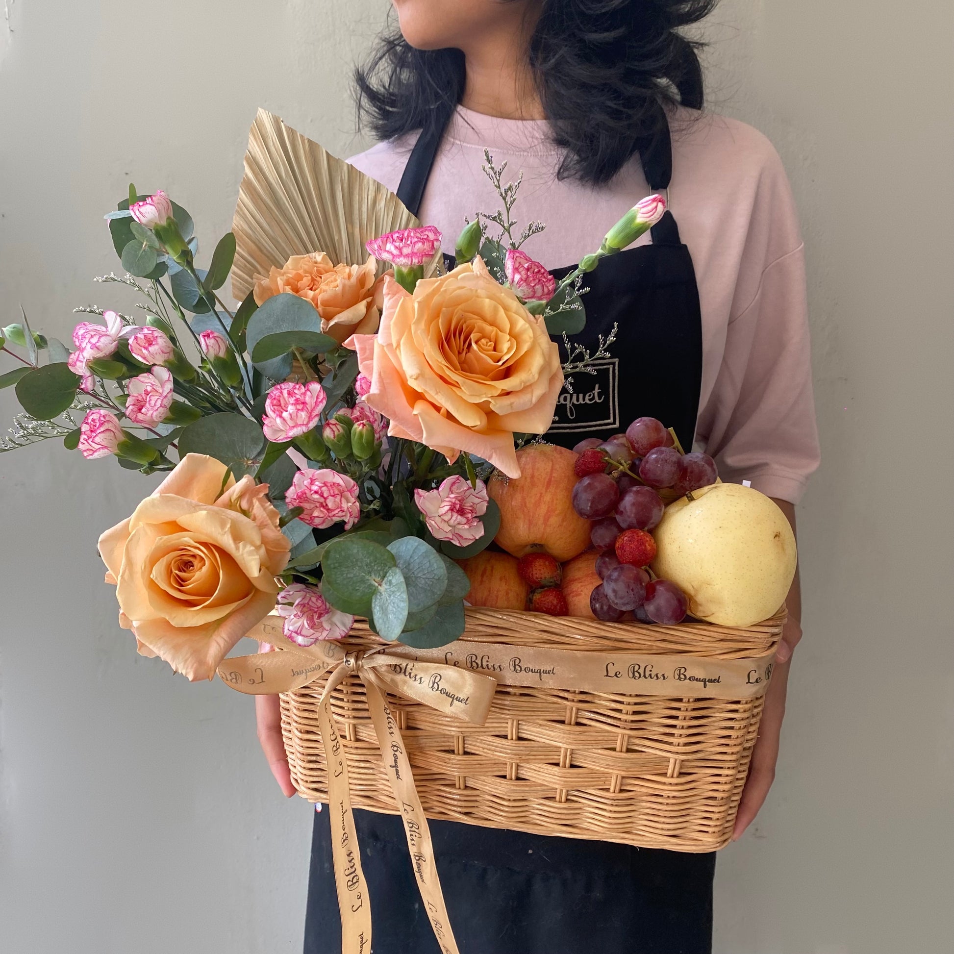 Keranjang anyaman cantik berisi buah-buahan segar dan bunga mawar peach yang memikat, dipegang oleh seorang wanita dengan kemeja merah muda. Tersedia untuk pengiriman di Jakarta, Bintaro, Bogor, Bekasi, dan Depok