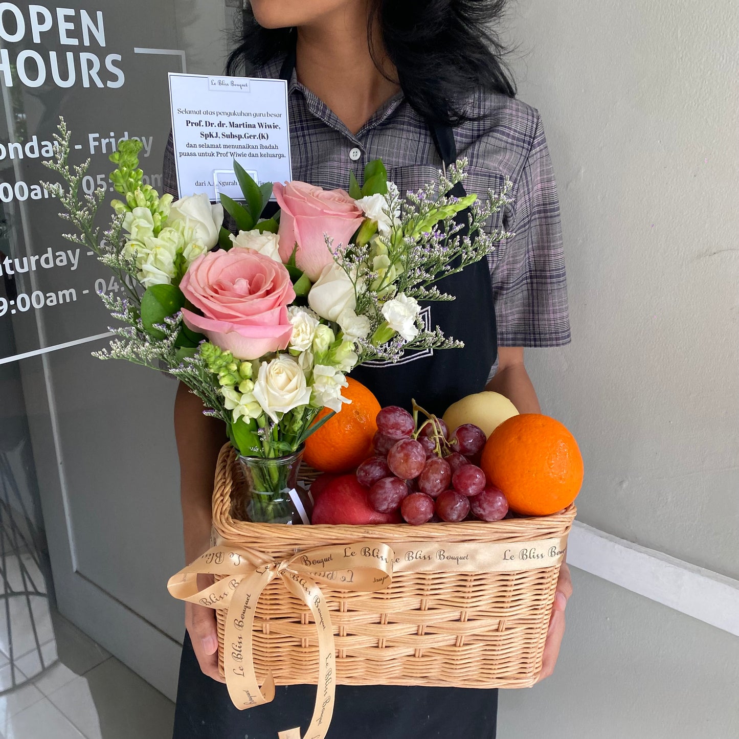A person holding a wicker fruit parcel containing an assortment of fresh fruits and roses, with a greeting card visible. Hermosa Fruit Parcel – Parcel buah dan bunga Hermosa pink premium dari Le Bliss Bouquet Surabaya, cocok untuk hadiah ulang tahun dan kado elegan. parcel buah surabaya