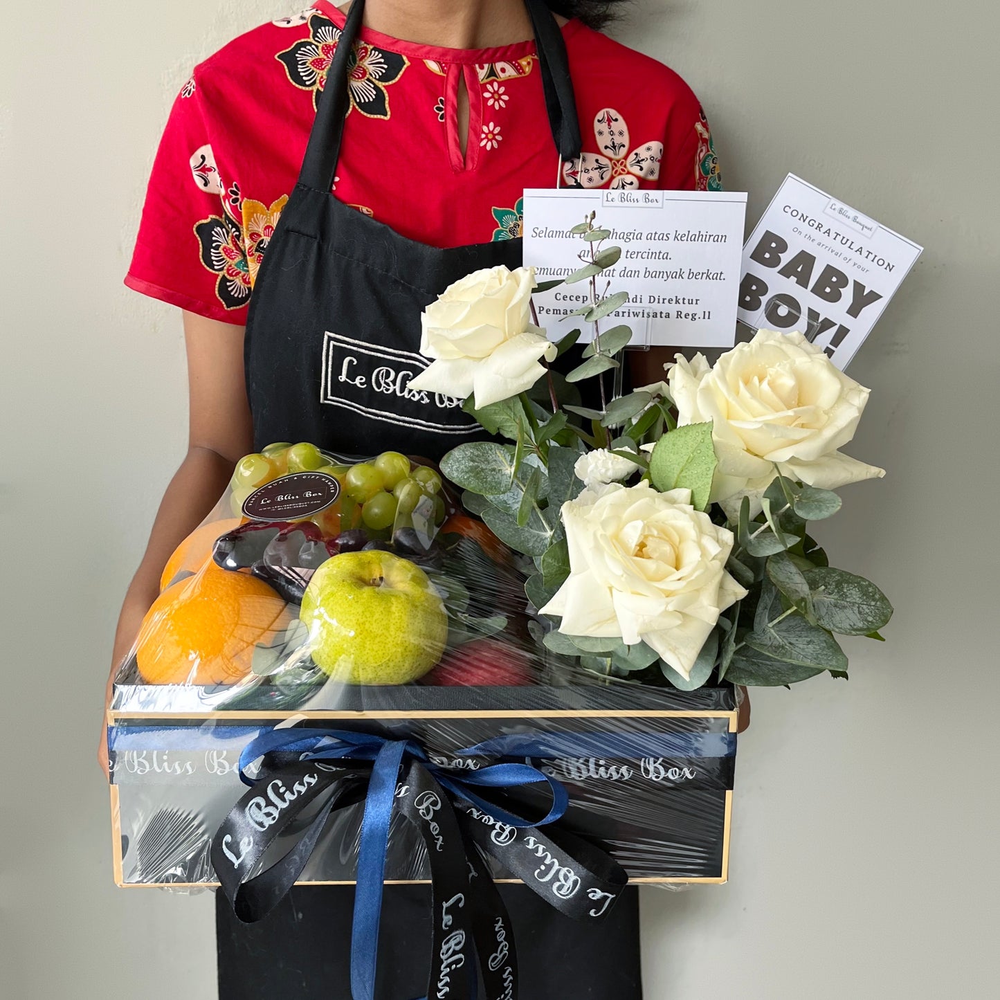 A person holding a gift box containing a selection of fruits and a bouquet of flowers, with a greeting card attached. parcel buah surabaya, parcel buah jakarta