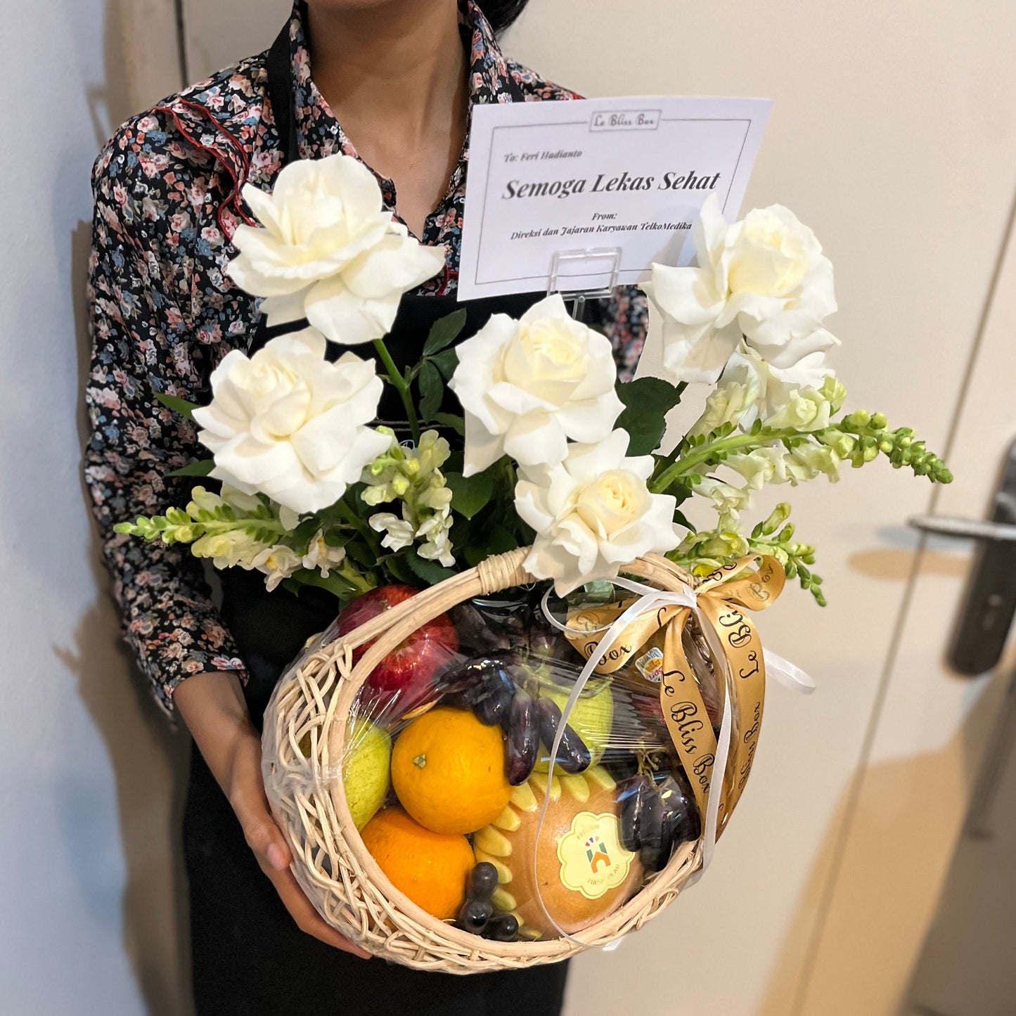 A person holding a gift basket containing fresh fruits, grapes, and a white roses. Fruit hamper jakarta, surabaya, cirebon by le bliss box