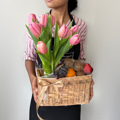 A person holding a wicker basket containing fresh tulips and assorted fruits with a customizable greeting card. Tulip Fruit Parcel – Parcel buah premium dengan bunga tulip segar dari Le Bliss Bouquet Surabaya, cocok untuk hadiah mewah dan hampers elegan. parcel buah surabaya