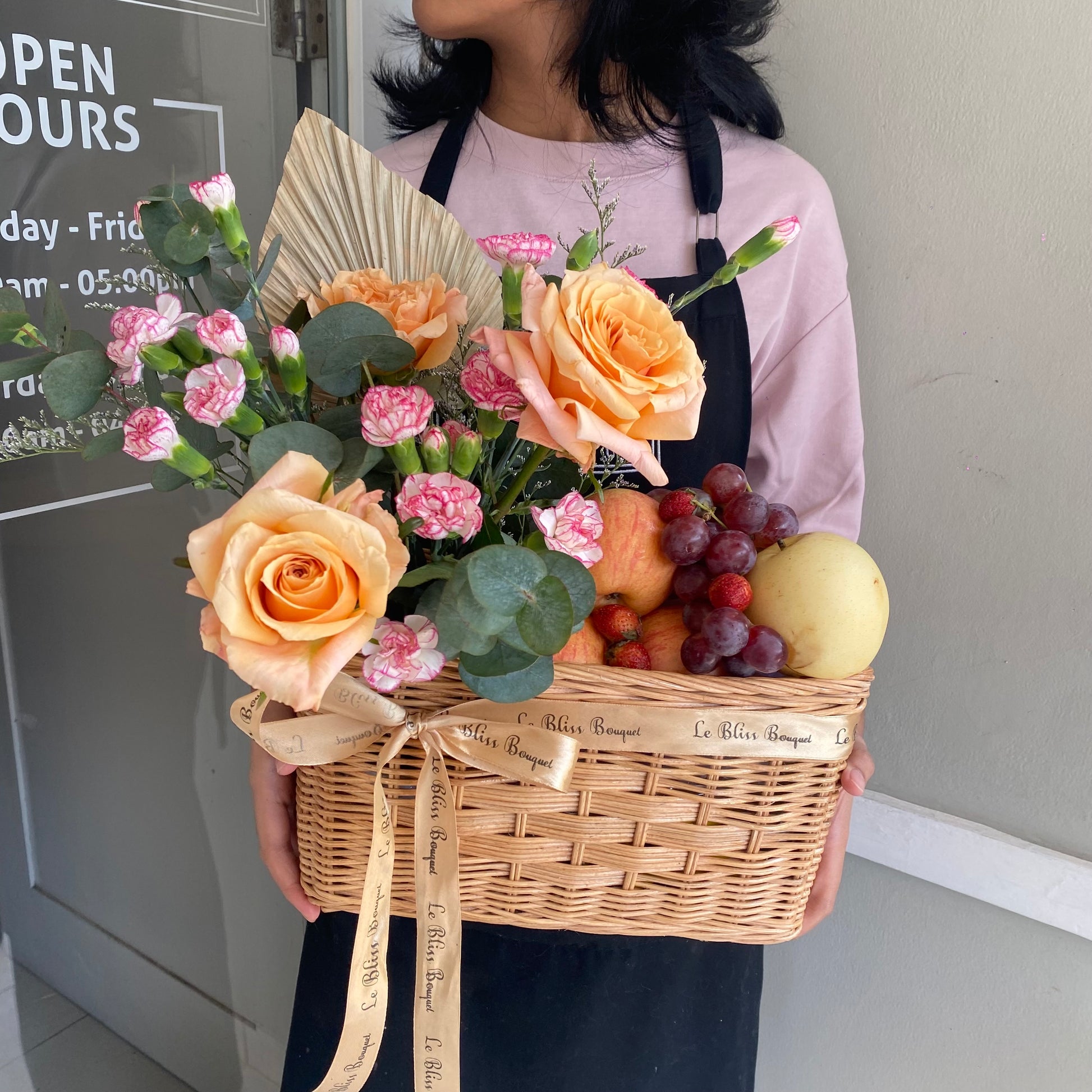 Keranjang anyaman cantik berisi buah-buahan segar dan bunga mawar peach yang memikat, dipegang oleh seorang wanita dengan kemeja merah muda. Tersedia untuk pengiriman di Jakarta, Bintaro, Bogor, Bekasi, dan Depo. parcel buah surabaya