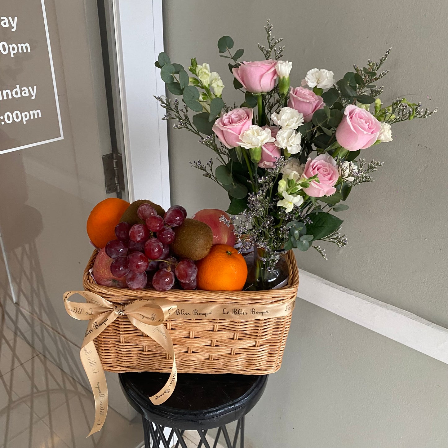 A wicker basket containing an assortment of fruits such as oranges, kiwi, grapes, and apples, accompanied by pink roses and white carnation flowers. fruit hamper surabaya, parcel buah surabaya