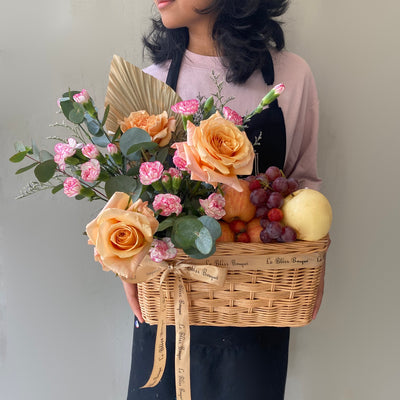 Keranjang anyaman cantik berisi buah-buahan segar dan bunga mawar peach yang memikat, dipegang oleh seorang wanita dengan kemeja merah muda. Tersedia untuk pengiriman di Jakarta, Bintaro, Bogor, Bekasi, dan Depok.