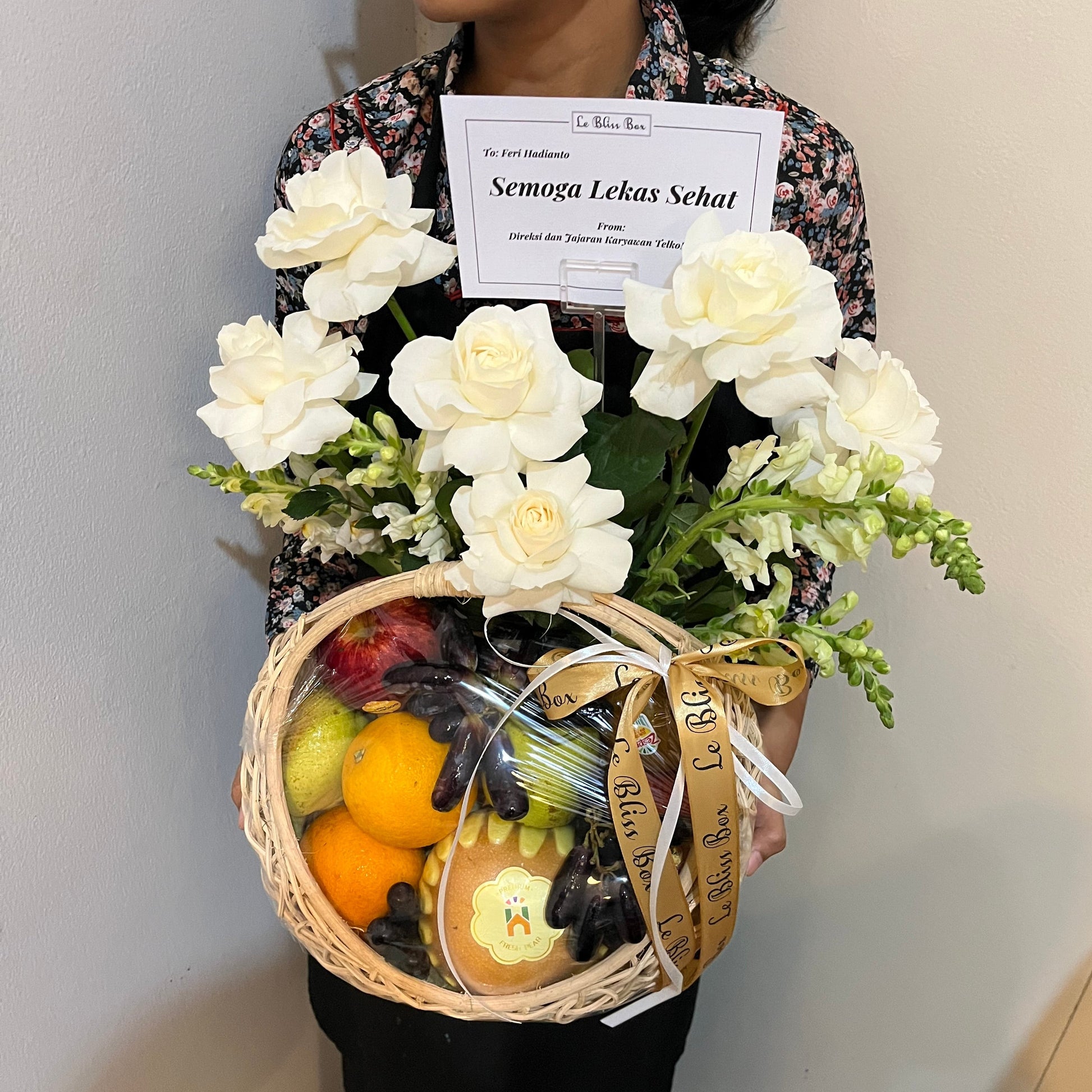 A person holding a gift basket containing fresh fruits, grapes, and a white roses. Fruit hamper jakarta, surabaya, cirebon by le bliss box