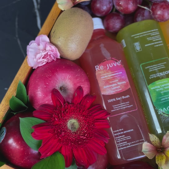 A wooden fruit box containing an assortment of fresh fruits such as apples, oranges, grapes, and bananas, along with two bottles of juice and decorative flowers. rejuve juice fruit hamper jakarta, surabaya, cirebon